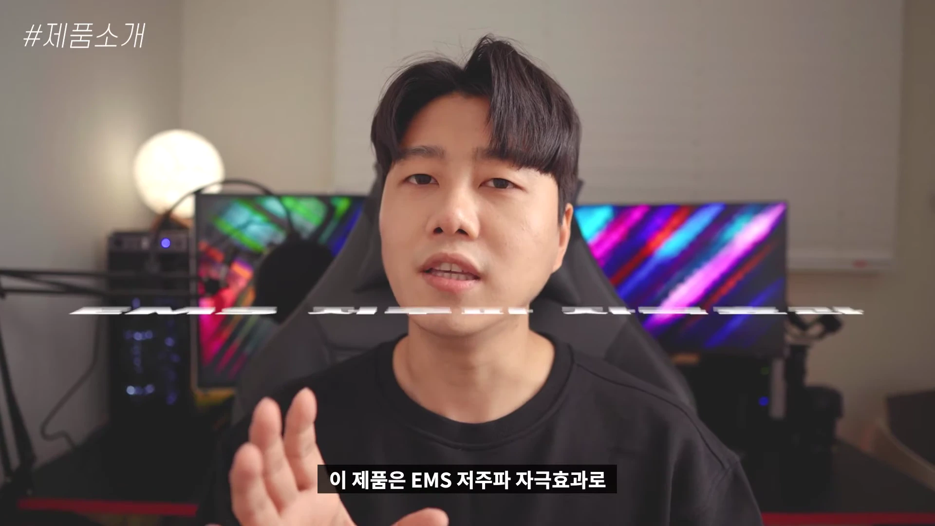 실내에서 검은색 상의를 입은 남성이 카메라를 보며 이야기하고 있으며, 배경에는 컴퓨터 모니터와 조명이 보입니다.