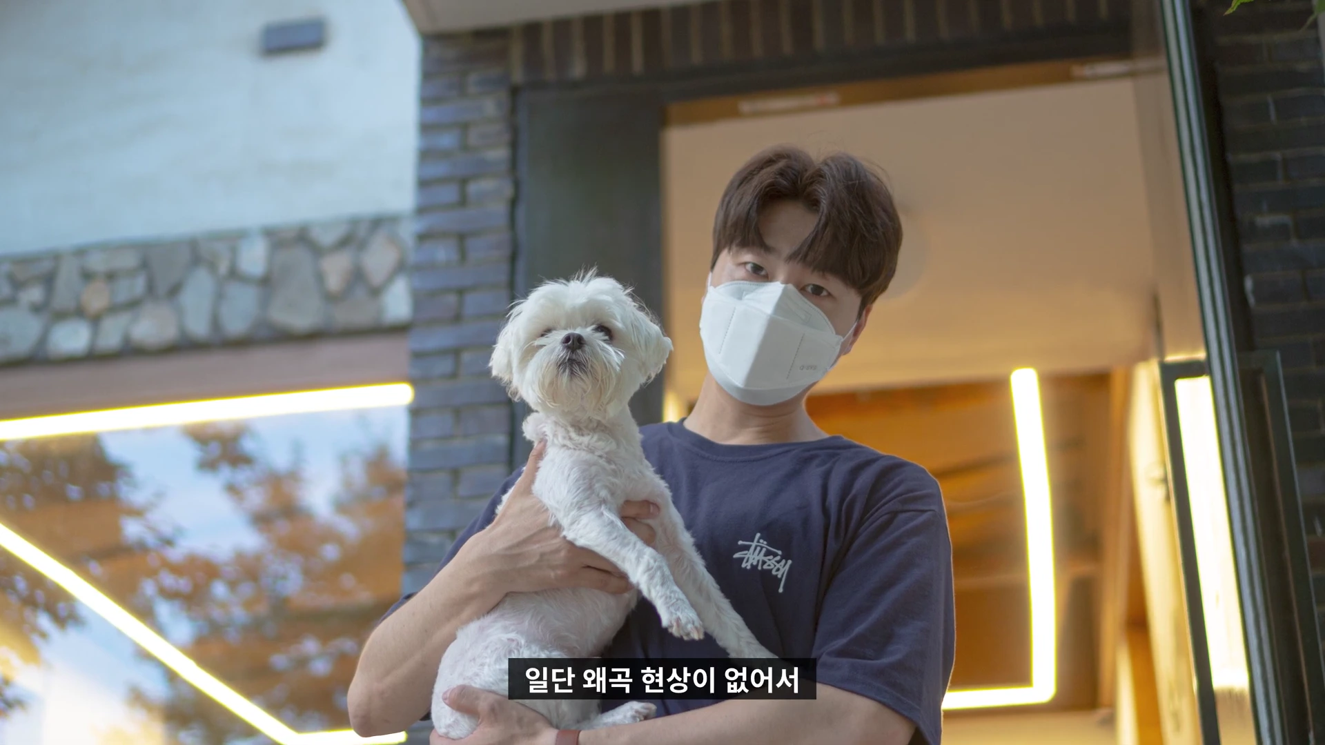 마스크를 쓴 남성이 야외에서 흰색 강아지를 두 손으로 안고 카메라를 응시하고 있으며 하단에 '일단 왜곡 현상이 없어서'라는 한국어 자막이 표시되어 있습니다.