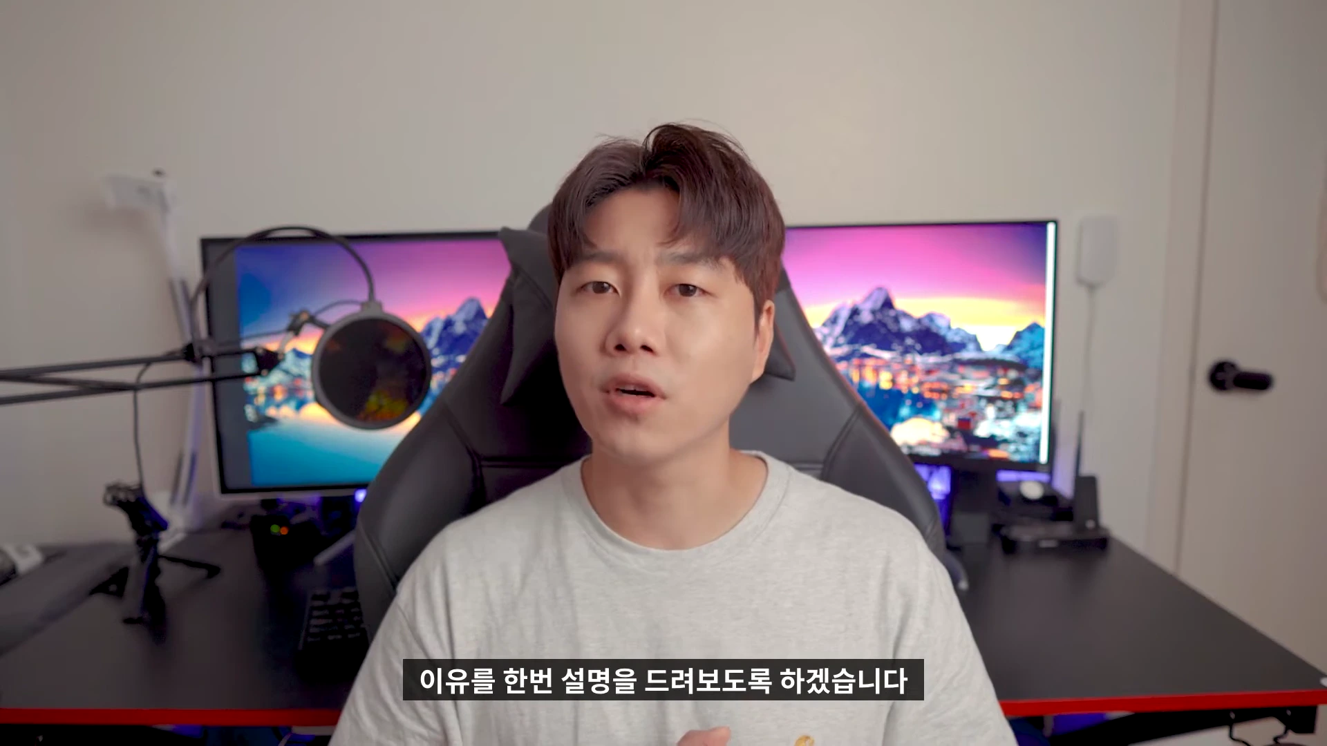 실내 책상 앞에 앉아 있는 남성이 카메라를 보며 이야기하고 있으며, 배경으로 모니터와 마이크가 보입니다.