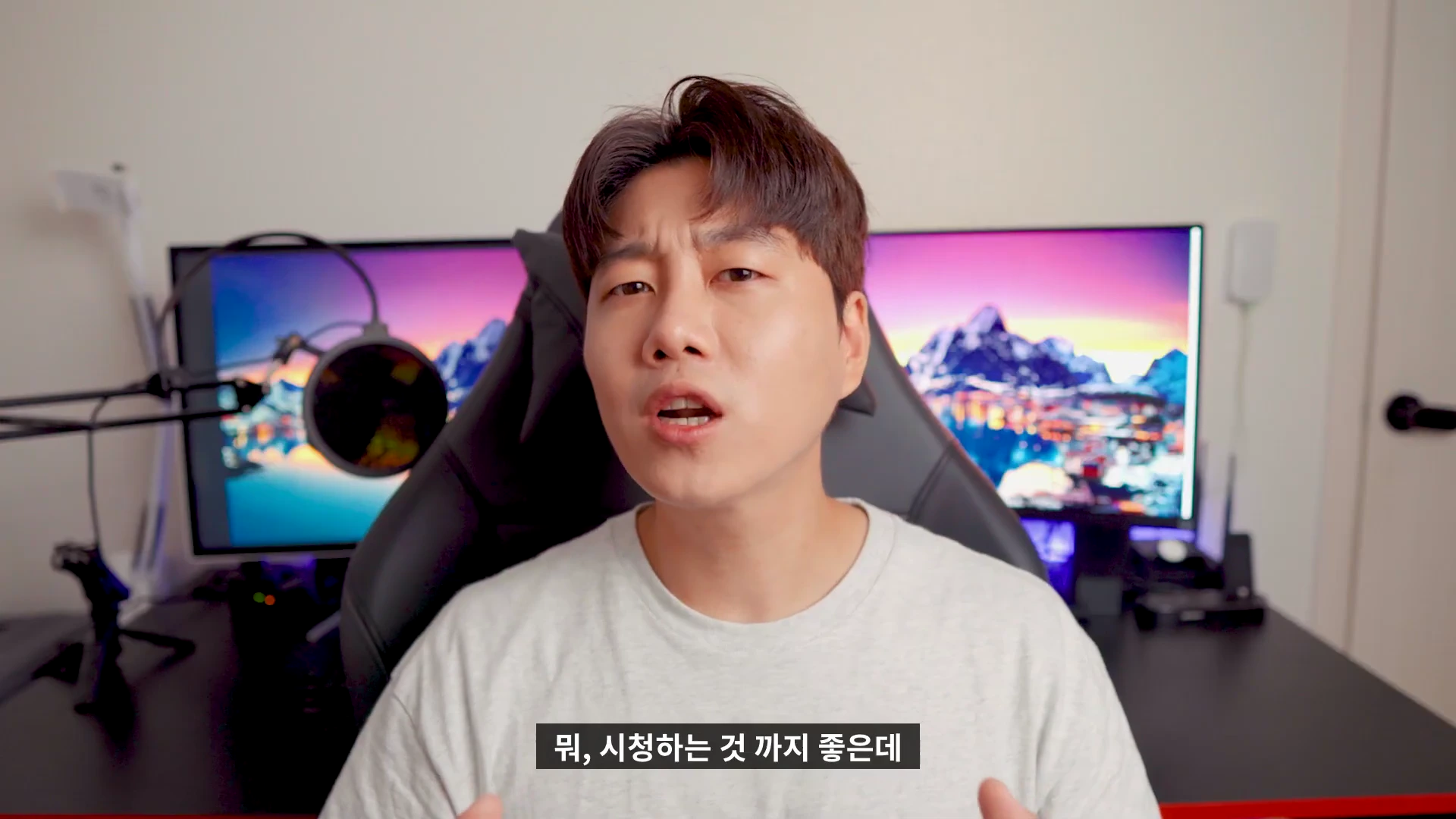 실내 책상에 앉아 카메라를 정면으로 바라보며 이야기하는 남성 뒤로 두 대의 모니터가 설치되어 있고, 화면에는 화려한 색감의 노을 풍경 이미지가 떠 있습니다.