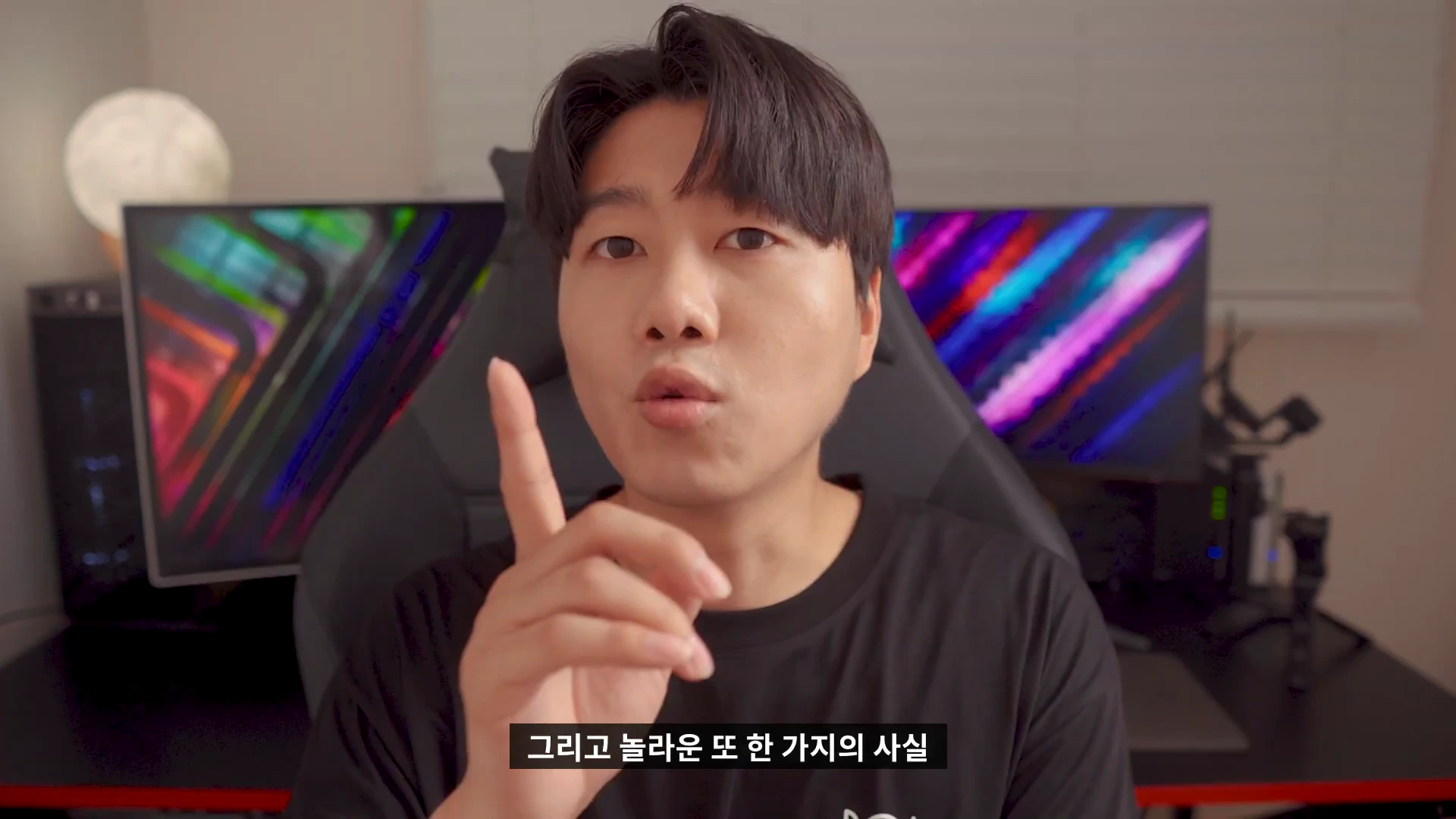 검은색 티셔츠를 입은 남성이 방 안에서 한 손가락을 들어 보이며 카메라를 정면으로 응시하고 있고 뒤쪽으로 조명이 켜진 모니터들이 보입니다.