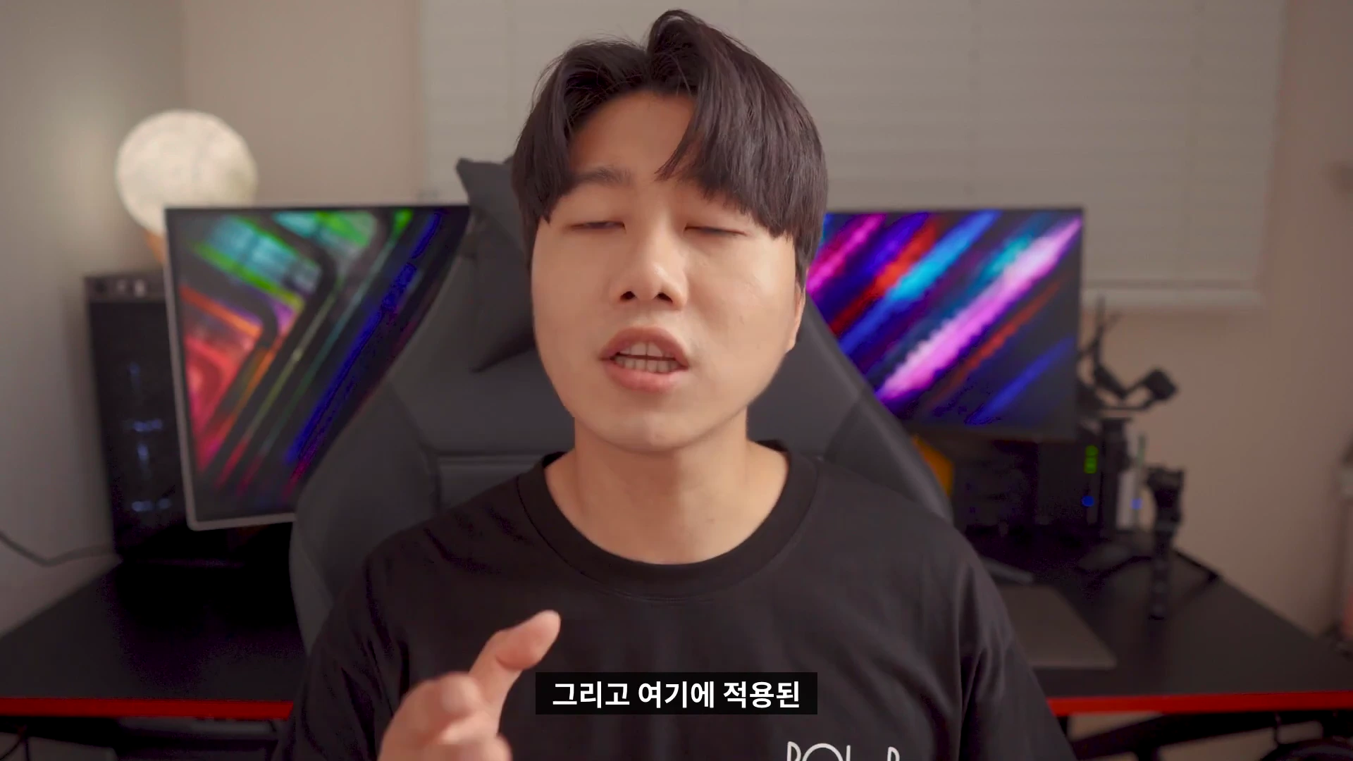실내에서 검은색 티셔츠를 입은 남성이 카메라를 보며 이야기를 하고 있으며 배경에는 게임용 모니터와 조명이 비치고 있습니다.