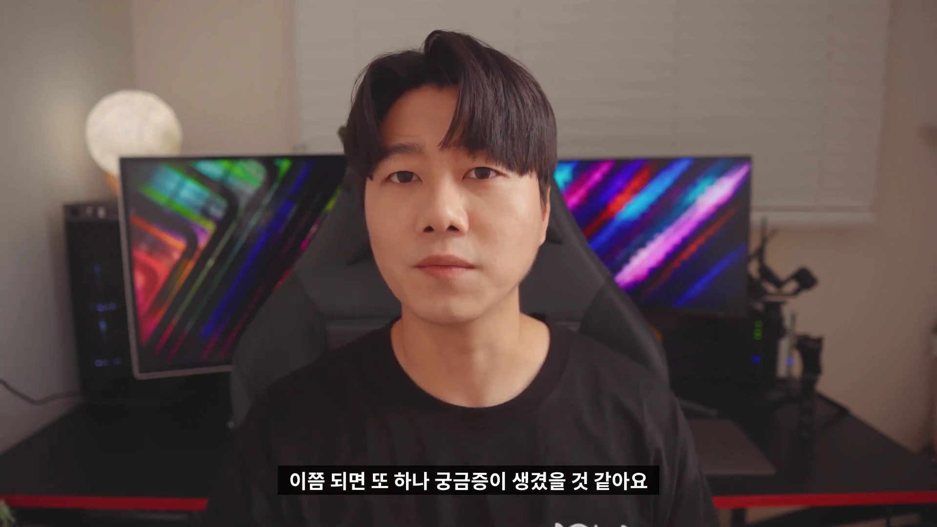 실내 스튜디오 의자에 앉아 정면을 응시하는 한 남성의 상반신 모습으로, 뒤편에 화려한 조명이 켜진 모니터 두 대가 배치되어 있습니다.