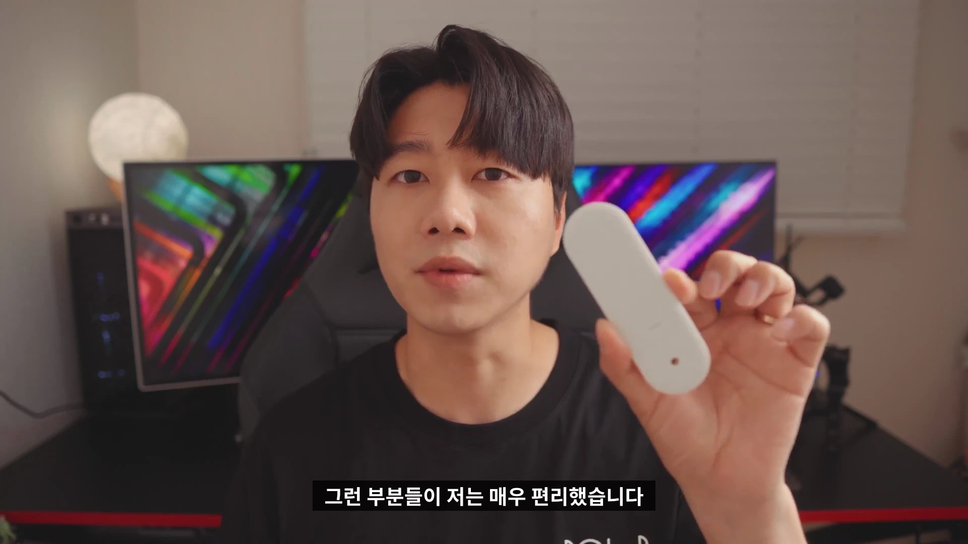 실내에서 검은색 티셔츠를 입은 남성이 카메라를 보며 흰색 리모컨을 들어 보이고 있다.