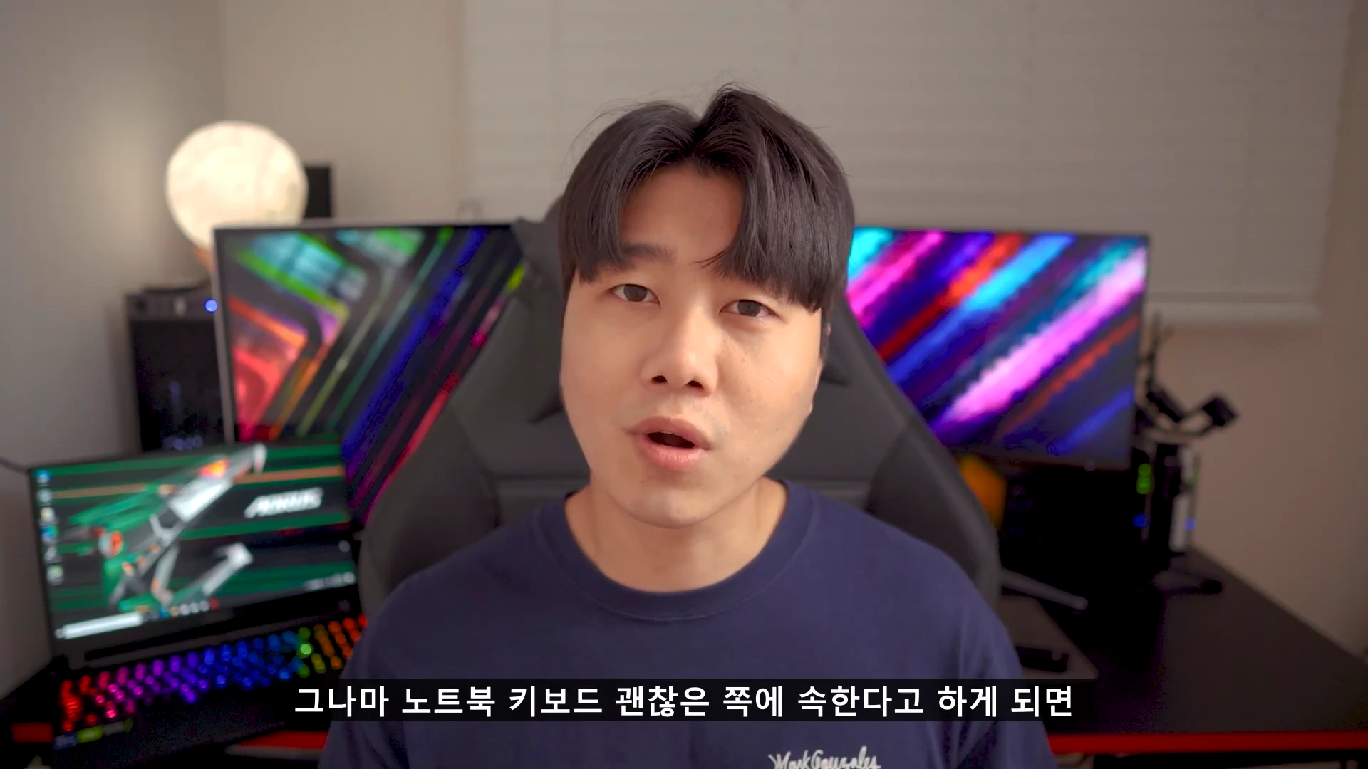 실내에서 게이밍 노트북과 모니터가 있는 책상 앞에 앉아 정면을 바라보며 이야기하는 남성 유튜버의 모습