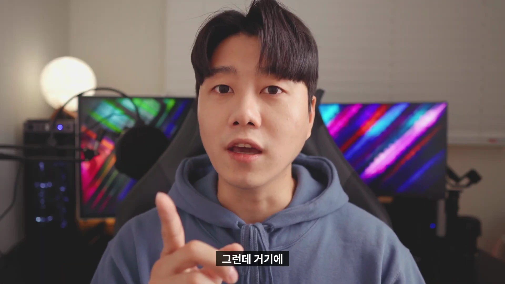 실내에서 후드티를 입은 남성이 카메라를 보며 손가락으로 가리키며 이야기하고 있으며, 배경에는 조명이 켜진 모니터들이 놓여 있습니다.