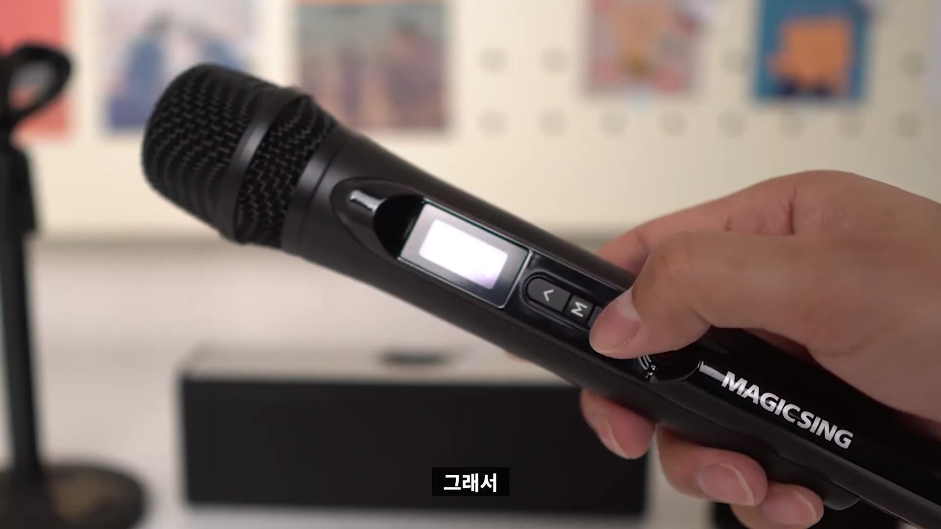 사용자의 손이 검은색 매직씽 블루투스 마이크의 버튼을 조작하고 있으며, 마이크 본체에 장착된 작은 LCD 화면과 조작 패널이 보입니다.