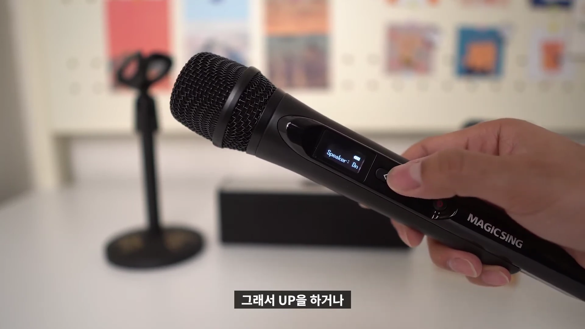 사람의 손이 검은색 매직씽 블루투스 마이크를 들고 있으며 마이크 중앙의 작은 디스플레이에는 'Speaker: Dn'이라는 설정값이 표시되어 있습니다. 배경에는 마이크 스탠드와 스피커가 흐릿하게 보입니다.