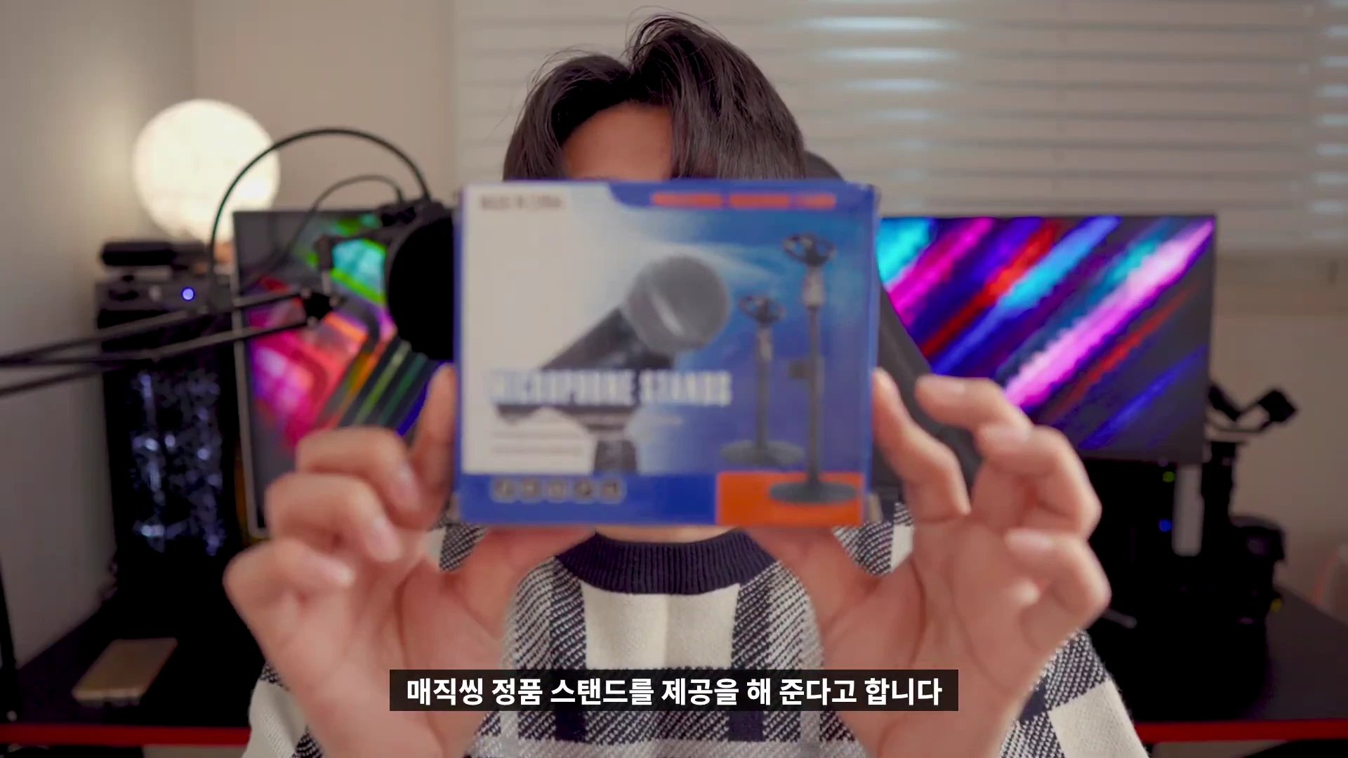 한 사람이 양손으로 마이크 스탠드 제품 상자를 들어 보이고 있으며, 배경에는 컴퓨터 모니터와 조명이 배치된 책상이 보입니다.