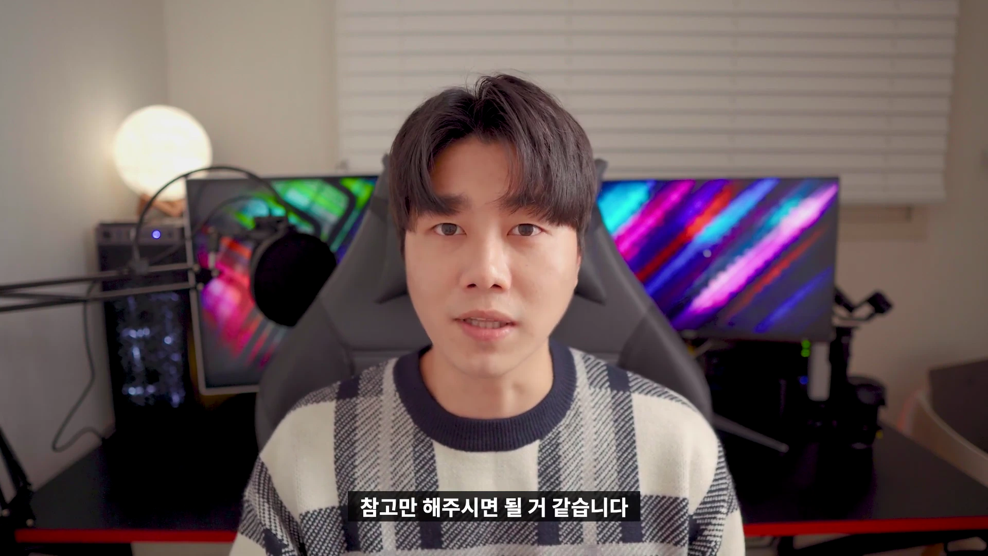 실내 책상에 앉아 있는 젊은 남성이 카메라를 정면으로 바라보며 이야기하고 있으며, 배경에는 컴퓨터 모니터와 조명이 배치되어 있습니다.