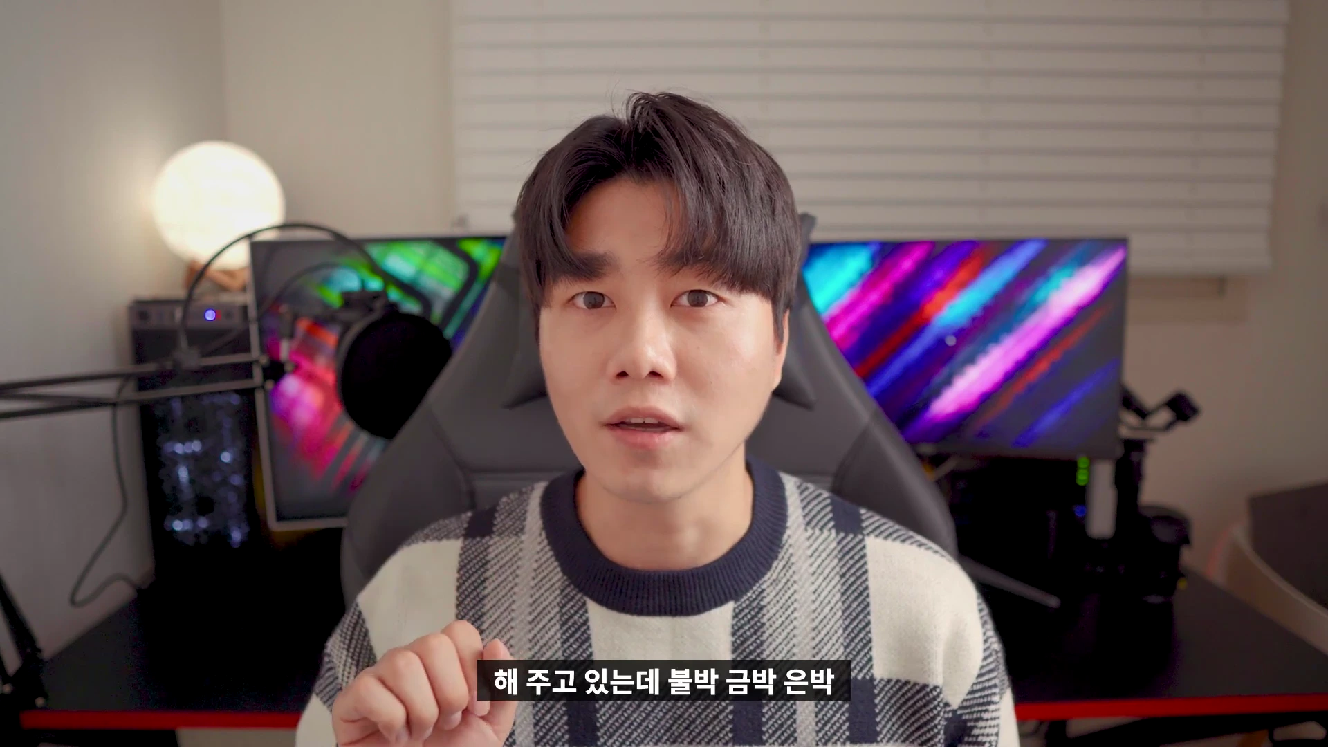 체크무늬 니트를 입은 남성이 카메라를 정면으로 응시하며 말을 하고 있는 모습입니다. 배경으로는 게이밍 의자와 조명이 켜진 모니터들이 보입니다.