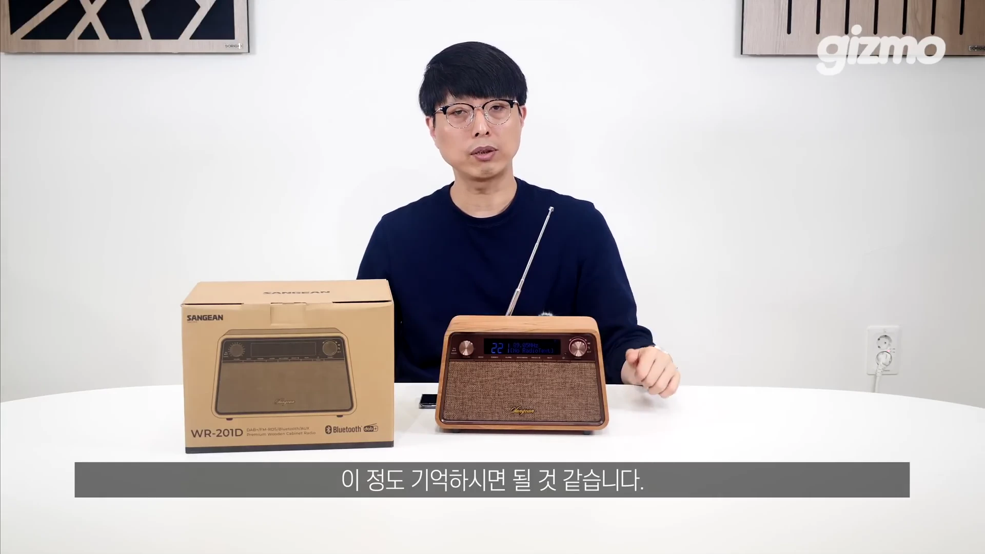 흰색 테이블 앞에 안경을 쓴 남성이 앉아 있고 그의 앞에는 제품 상자와 우드 케이스로 마감된 라디오가 놓여 있습니다.