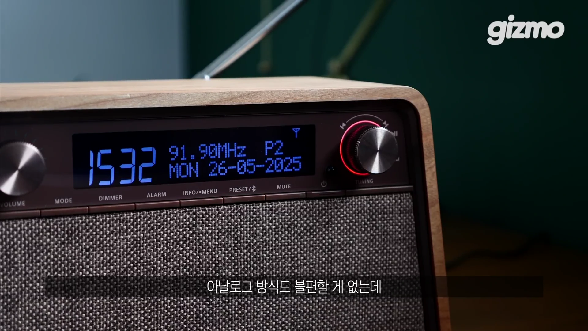나무 재질 프레임과 직물 스피커 그릴이 특징인 탁상형 라디오의 전면부로, 파란색 글씨가 표시되는 디지털 디스플레이와 조작 다이얼이 보입니다.