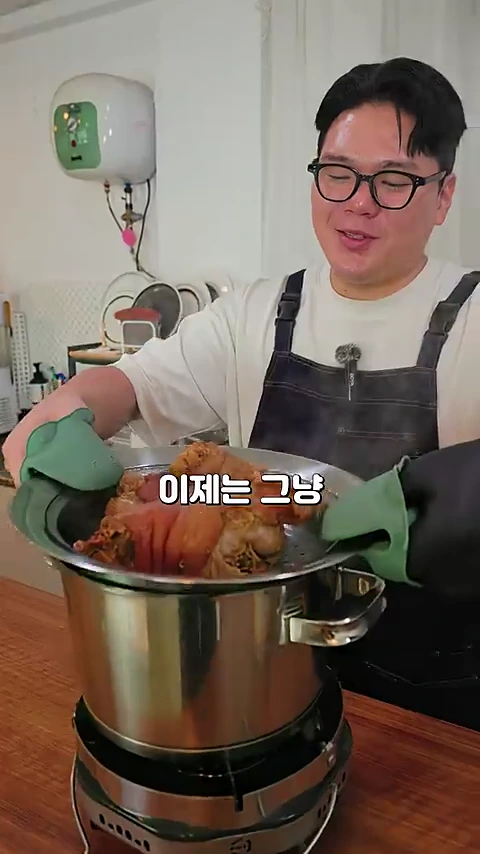 안경을 쓴 남성이 앞치마를 입고 초록색 실리콘 오븐 장갑을 낀 채 찜기에 담긴 족발을 들어 올리고 있는 모습