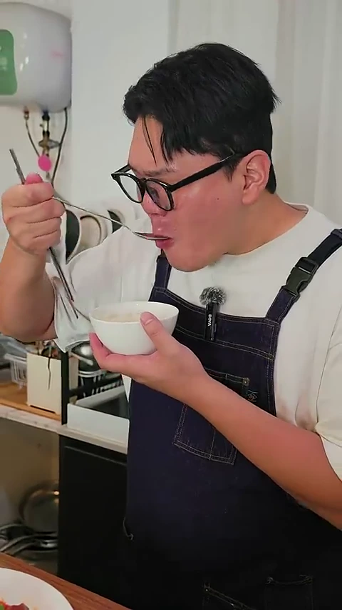 흰색 티셔츠와 남색 앞치마를 입고 뿔테 안경을 쓴 남성이 숟가락으로 음식을 떠서 먹고 있는 모습입니다.