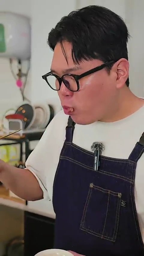 뿔테 안경을 쓴 남성이 앞치마를 두르고 젓가락으로 족발을 집어 입으로 가져가 맛보고 있다.