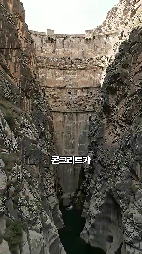 좁고 깊은 암벽 사이로 거대한 돌로 쌓아 올린 아치형 댐 벽면이 보이며, 화면 중앙 하단에 '콘크리트가'라는 자막이 겹쳐 있다.