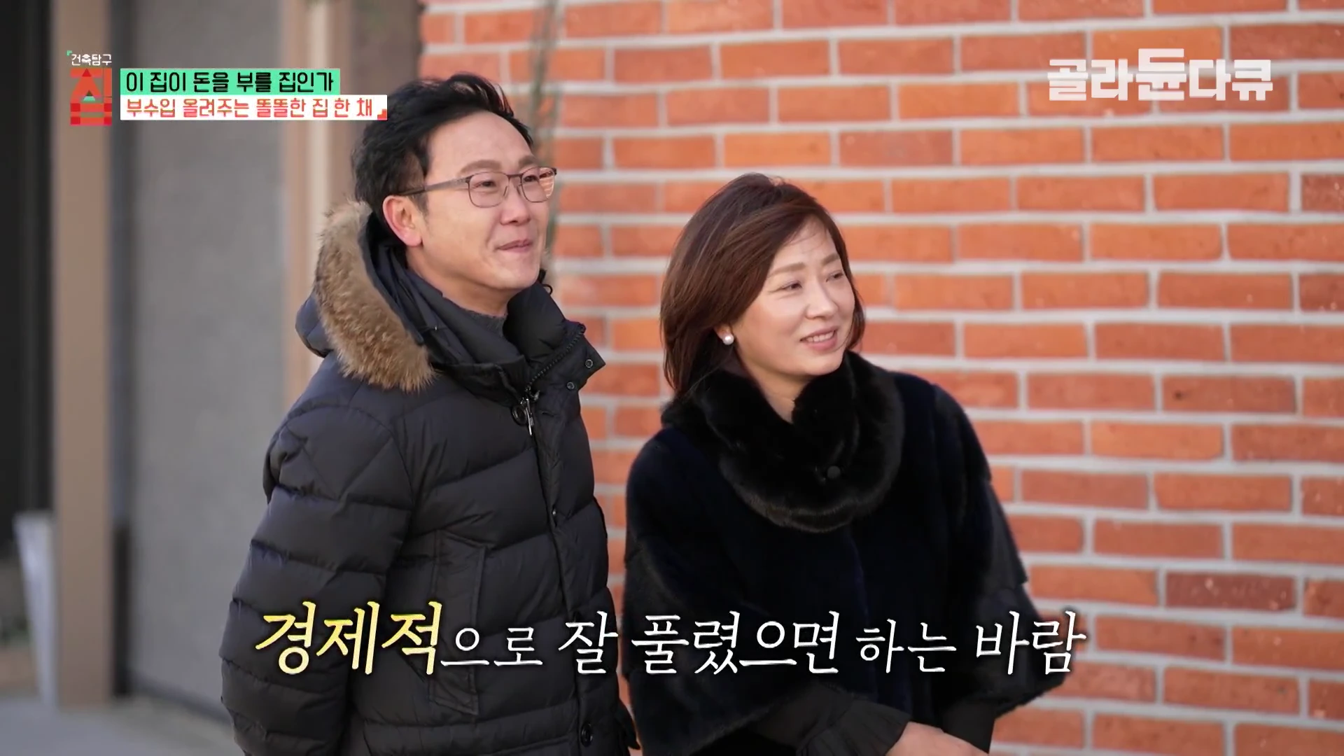 붉은 벽돌 벽을 배경으로 나란히 서서 미소 짓고 있는 중년 부부의 모습이 담긴 방송 화면