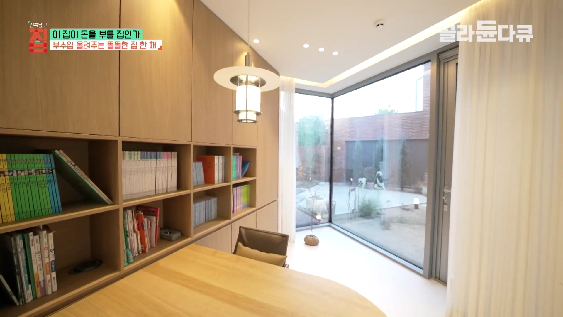 나무 소재의 붙박이 책장과 책상이 놓인 서재 공간으로, 모서리까지 이어진 통창 너머로 정원이 보이는 실내 모습.