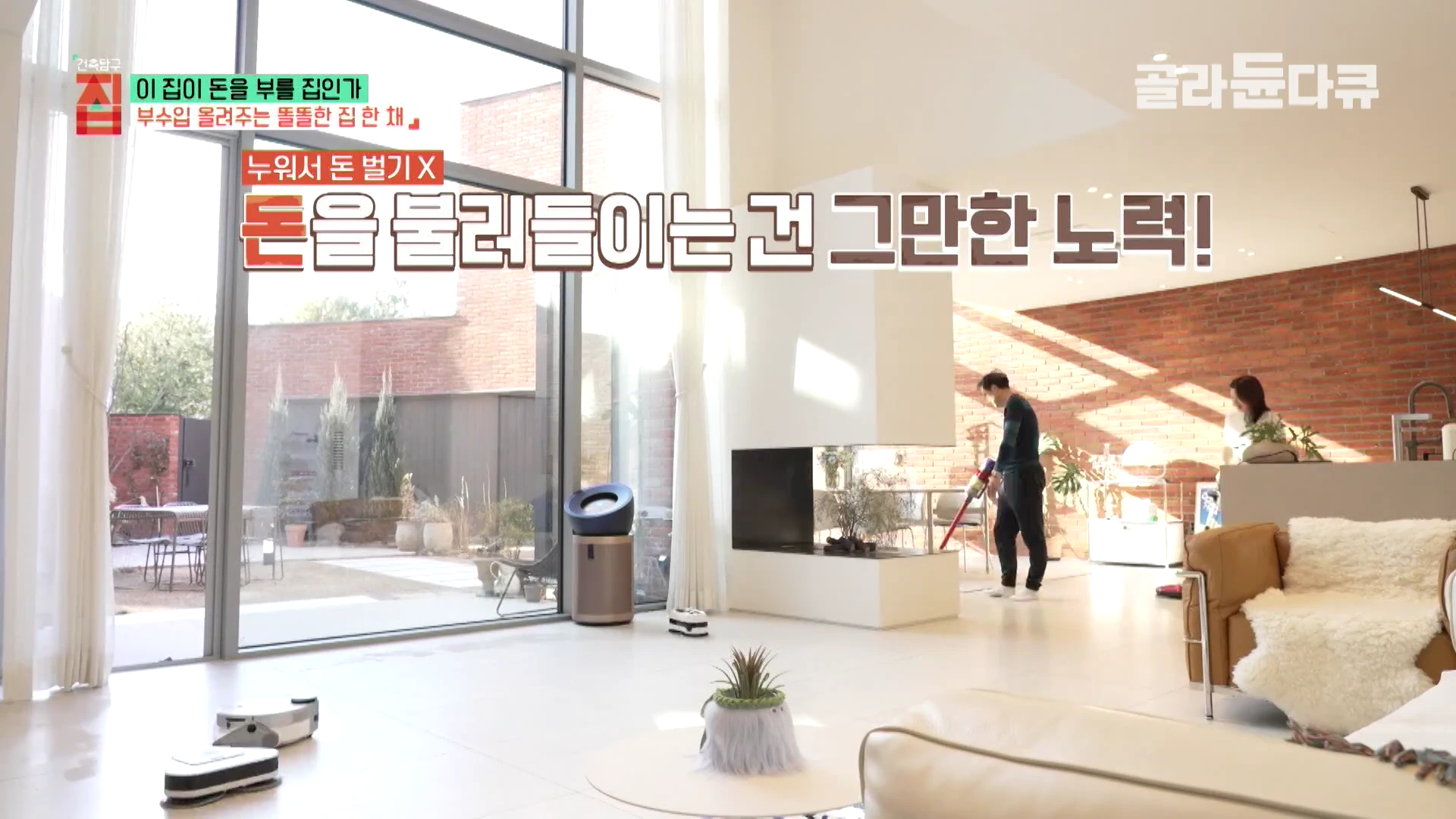 햇살이 가득 들어오는 높은 통창이 있는 거실에서 한 남성이 무선 청소기를 사용하고 있고, 바닥에는 로봇 청소기들이 다니고 있는 모습