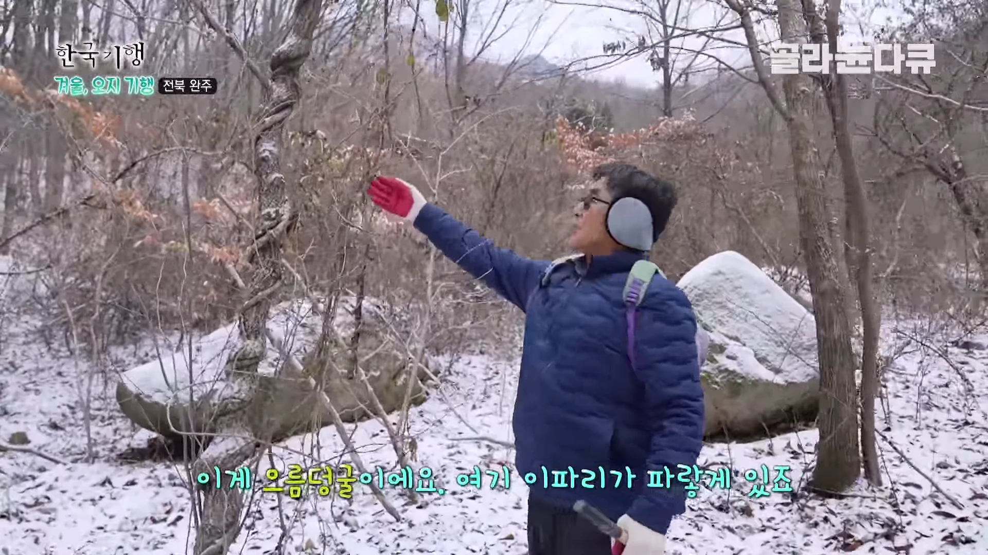눈 덮인 산길에서 파란색 패딩 점퍼를 입고 귀마개를 한 남성이 나무에 엉킨 덩굴을 가리키며 설명하고 있다.