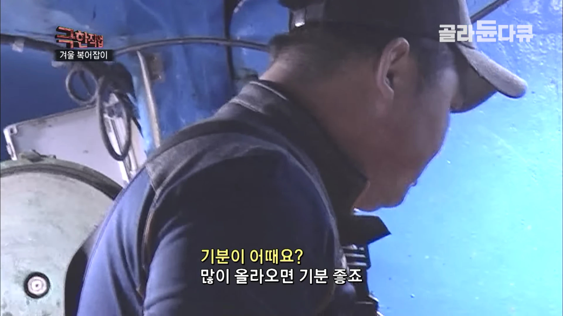 선상에서 모자를 쓴 어부가 작업에 열중하고 있는 모습을 담은 다큐멘터리 영상 화면입니다. 화면 중앙에는 '기분이 어때요? 많이 올라오면 기분 좋죠'라는 자막이 노란색으로 표시되어 있습니다.