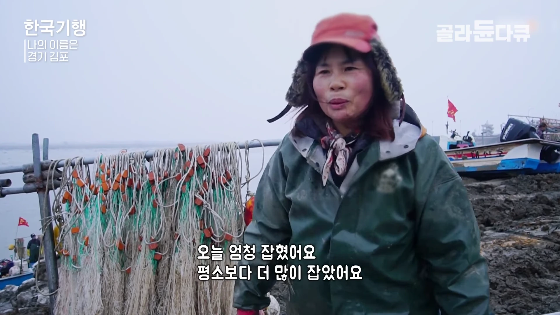 방한 모자를 쓰고 낚시용 녹색 점퍼를 입은 한 여성이 강가 그물 앞에서 인터뷰하고 있는 모습