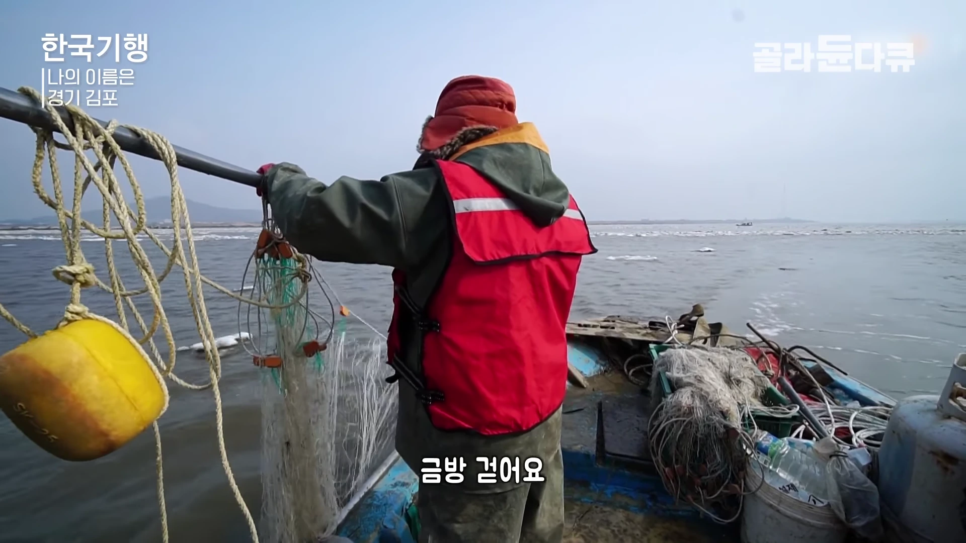 두꺼운 방한복과 빨간 조끼를 입은 어부가 유빙이 떠다니는 한강 하구에서 배 위에 서서 그물을 걷어 올리고 있는 모습