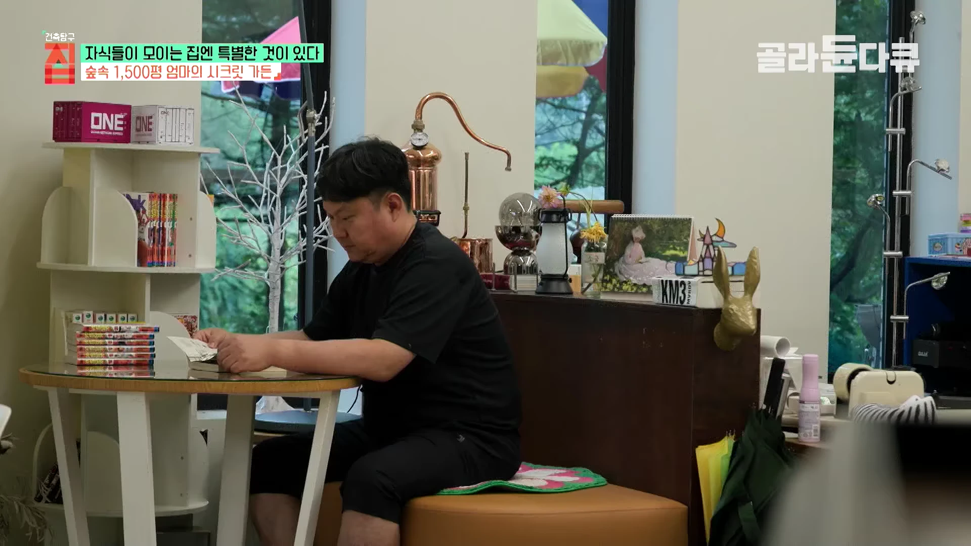 실내에서 검은 옷을 입은 남성이 원형 테이블에 앉아 책을 읽고 있는 모습과 뒤편의 큰 창문을 통해 밖의 수풀이 보이는 실내 공간
