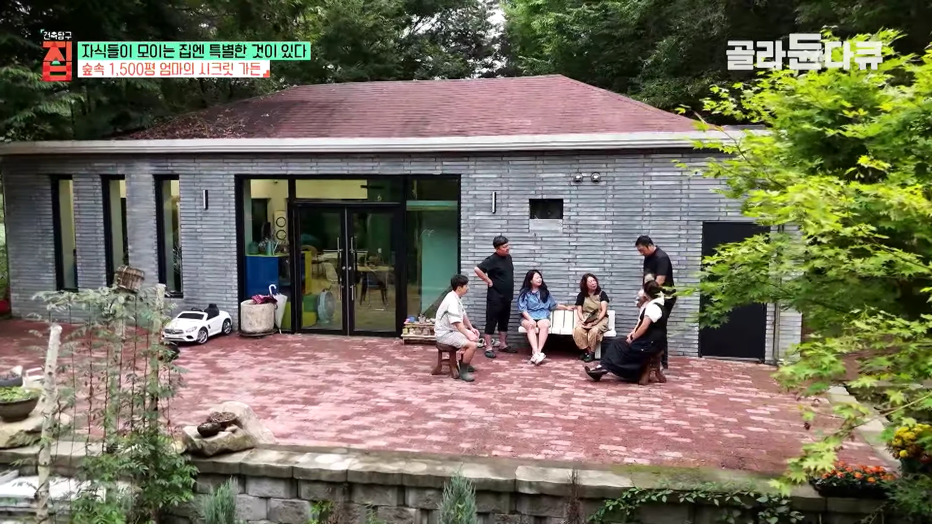 숲속에 자리 잡은 회색 벽돌 단층 건물 앞에 다섯 명의 가족이 야외 테라스에 앉아 대화를 나누고 있는 모습이다.