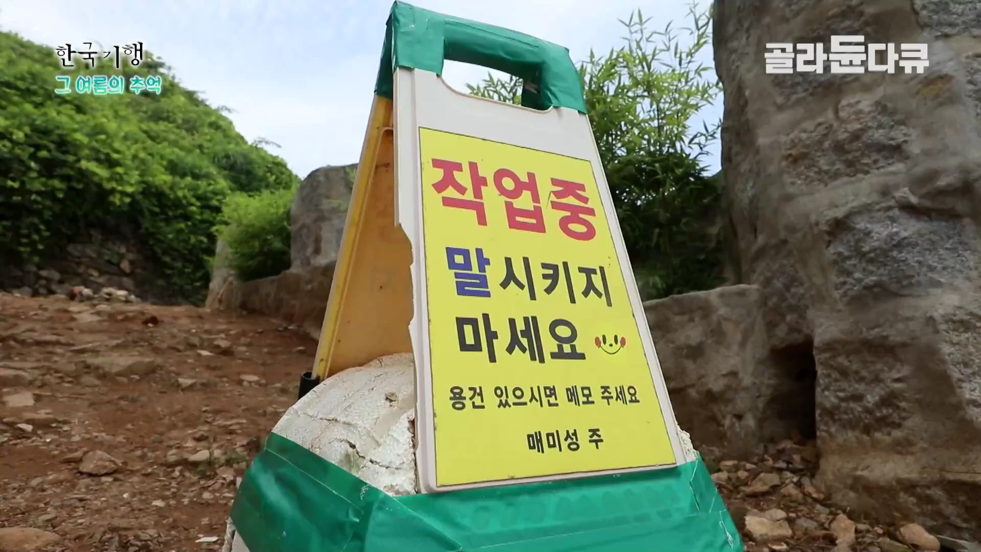 돌길 위에 '작업중 말시키지 마세요'라고 적힌 노란색 접이식 안내판이 놓여 있고, 배경으로 흙길과 돌벽이 보입니다.