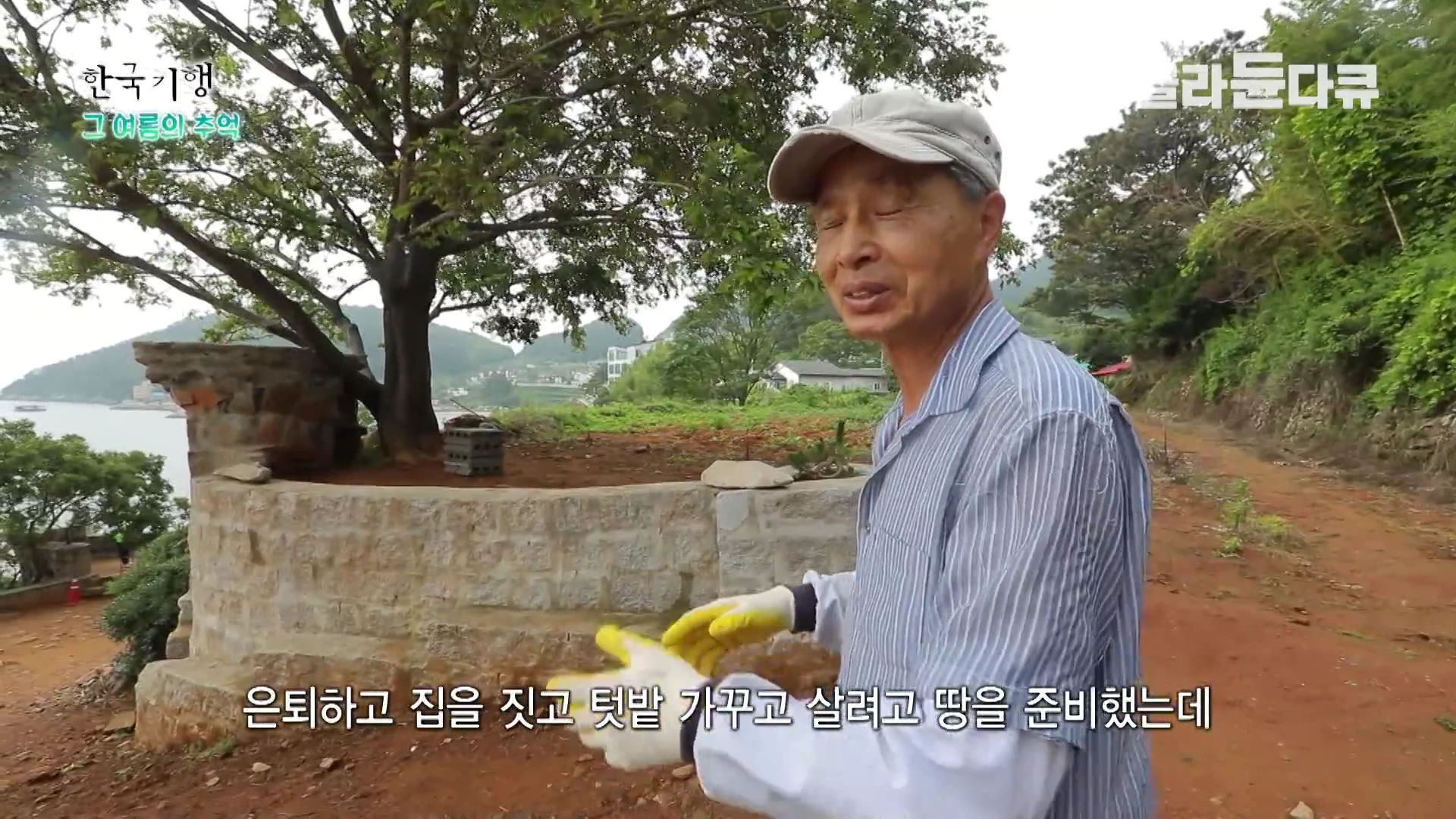 모자를 쓰고 작업용 장갑을 낀 한 남성이 야외에서 바닷가 근처의 돌 축대를 배경으로 서서 손짓하며 이야기를 나누고 있다.