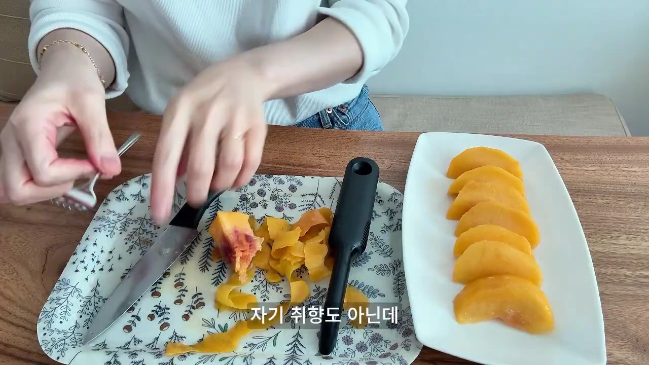 나무 식탁 위에 놓인 꽃무늬 쟁반 위에서 한 사람이 칼을 사용해 복숭아 껍질을 벗기고 있으며, 옆에는 껍질을 벗긴 복숭아 조각들이 접시에 정갈하게 담겨 있습니다.