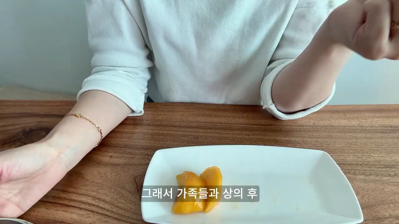 흰 긴소매 옷을 입은 사람이 나무 식탁 위에 놓인 접시에 잘라 둔 복숭아 조각을 손으로 가리키고 있다.