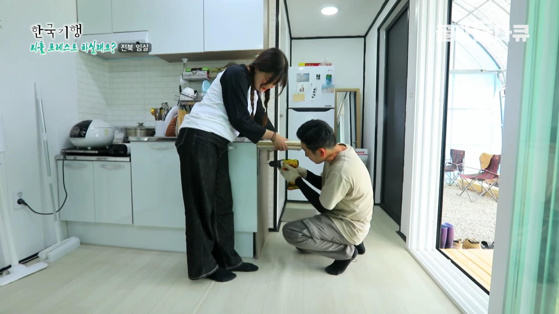 실내 컨테이너 주택 주방에서 한 남성이 전동 드릴로 선반을 설치하고 있고, 한 여성이 옆에서 이를 잡아주고 있는 모습
