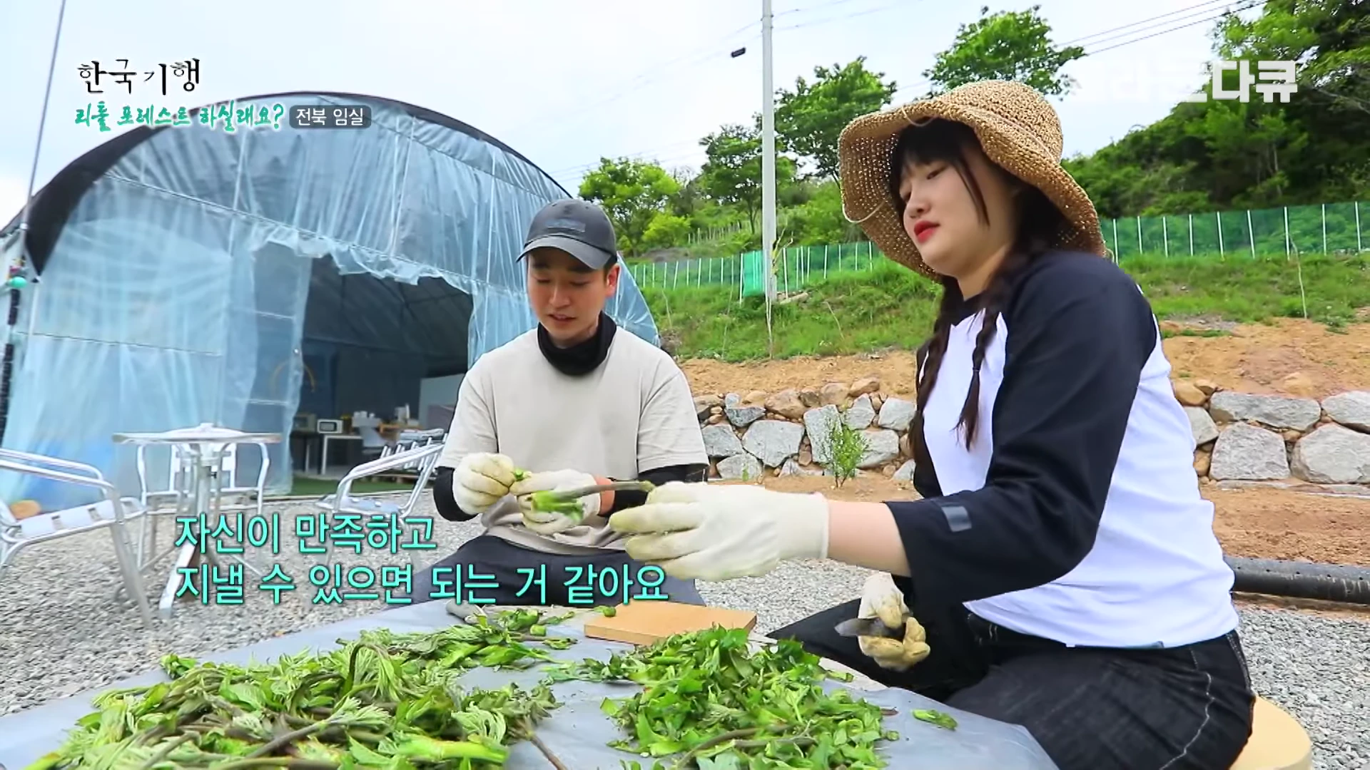 한 쌍의 부부가 밀짚모자와 작업 장갑을 착용하고 비닐하우스 앞 야외 테이블에 앉아 함께 채소를 다듬고 있는 모습