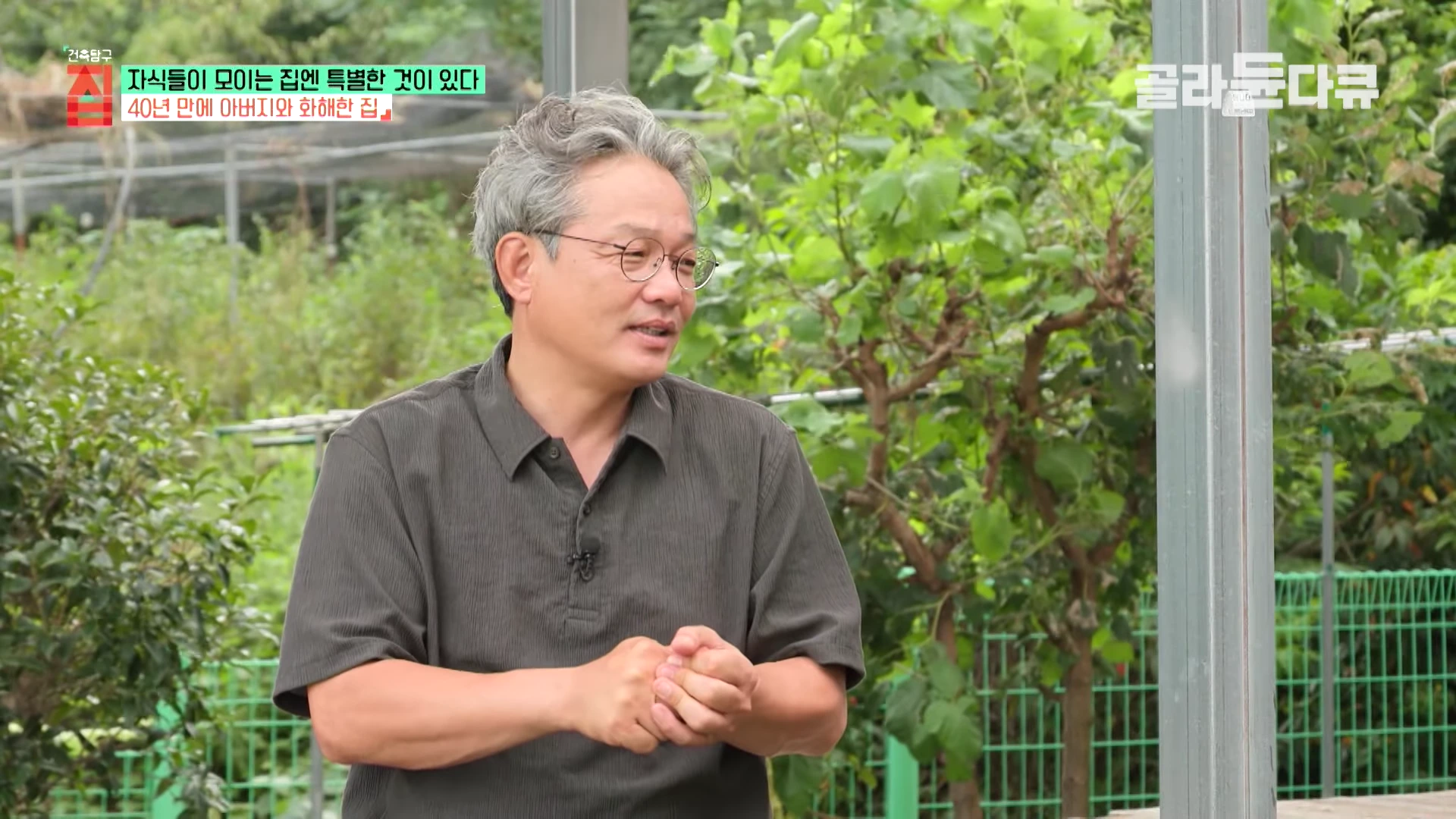 흰 머리에 안경을 쓴 중년 남성이 야외 정원 배경에서 대화를 나누며 손을 모으고 있다.
