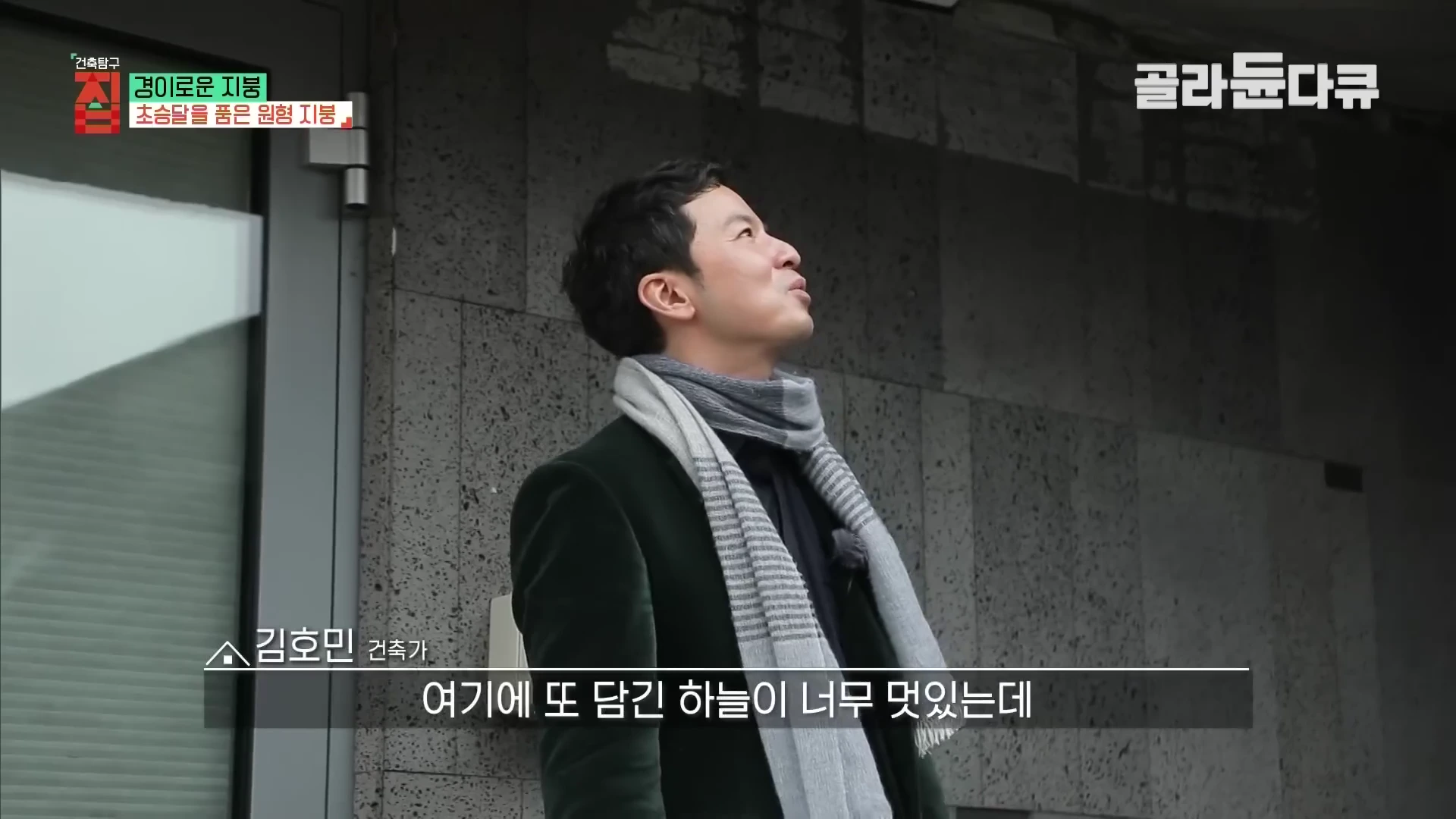 짙은 녹색 코트를 입은 한 남성이 회색 현무암 판석으로 마감된 외벽 옆에서 고개를 들어 위쪽을 바라보고 있다. 화면 상단에는 방송 프로그램 로고가 표시되어 있다.