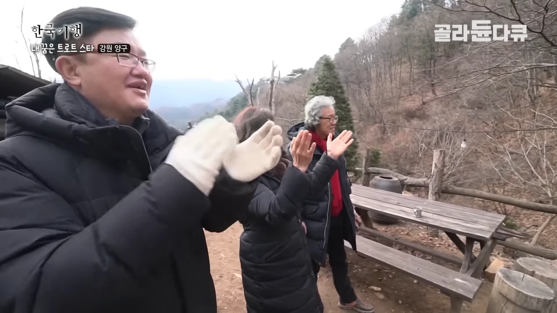 세 사람이 산골 야외에서 겨울 외투를 입고 밝게 웃으며 박수를 치고 있다.