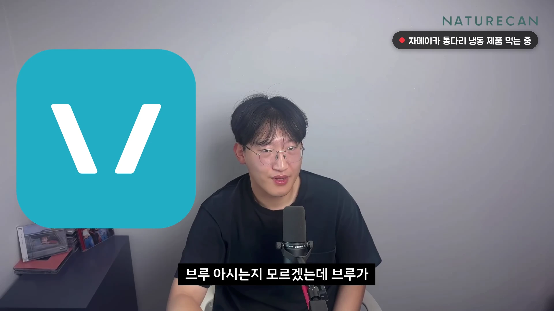 안경을 쓴 남성이 마이크 앞에서 이야기하고 있으며, 화면 왼쪽에는 민트색 배경에 흰색 'V'자가 그려진 로고가 있고, 오른쪽 상단에는 '자메이카 통다리 냉동 제품 먹는 중'이라는 텍스트 박스가 떠 있다.