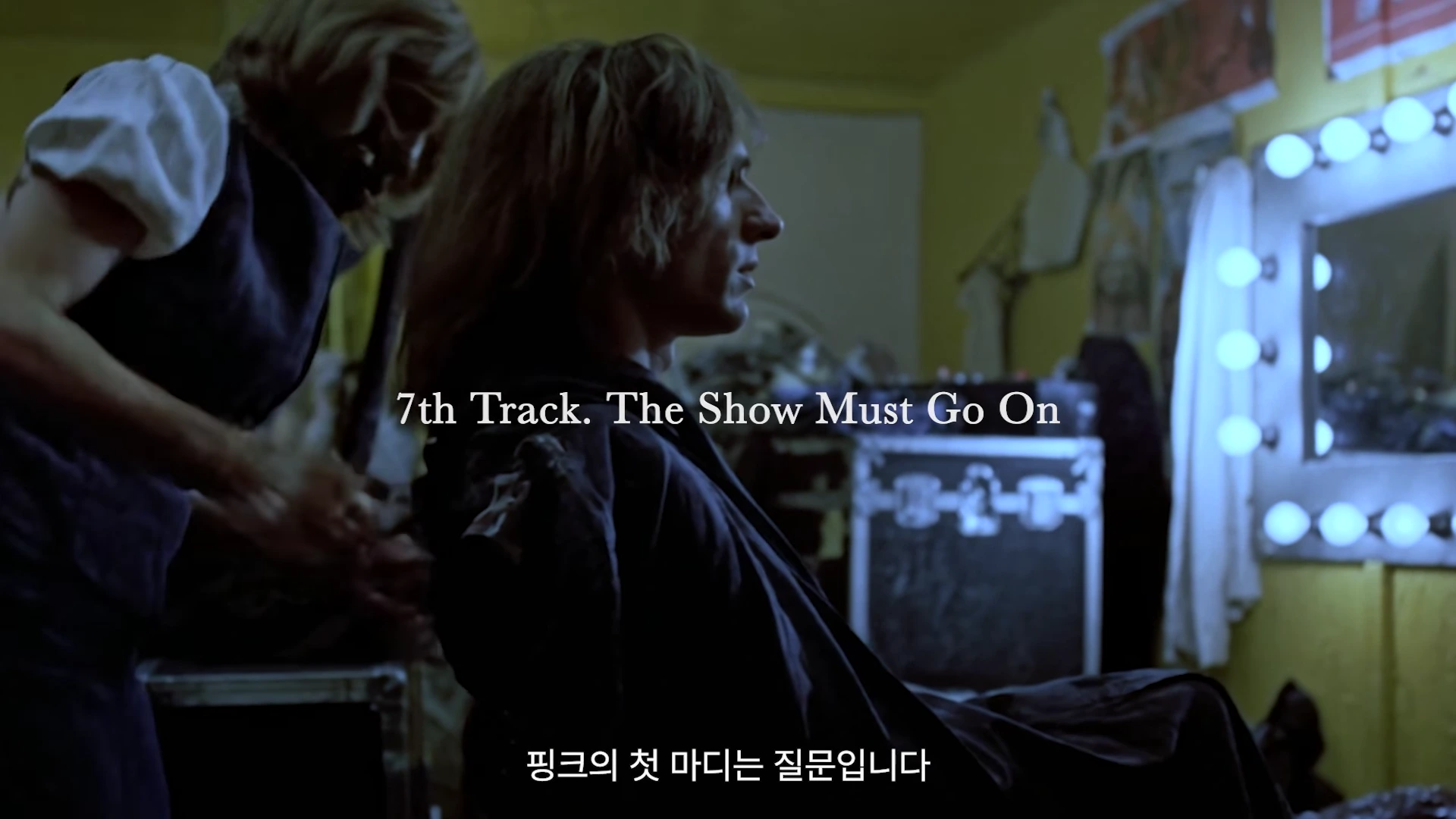 분장실에서 거울 조명을 마주하고 앉아 있는 긴 머리의 남성과 그의 뒤에서 머리를 만져주는 사람의 모습이 담긴 영화의 한 장면.