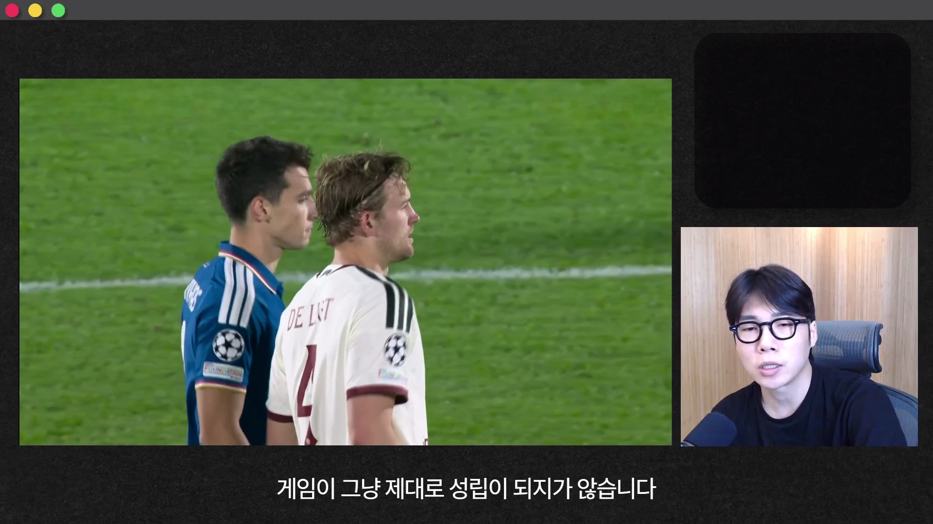 축구 경기 중 등 돌리고 서 있는 두 선수와 화면 오른쪽 하단에서 말하는 안경 쓴 남성을 담은 영상 화면