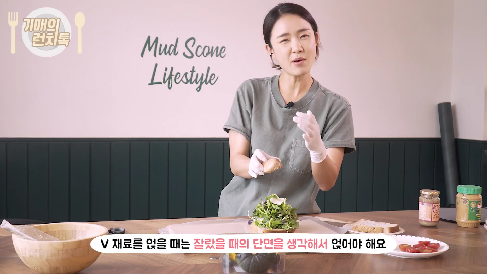 한 여성이 요리용 장갑을 끼고 샌드위치 재료를 손질하며 이야기를 나누고 있는 모습으로 책상 위에는 채소와 빵, 소스 등이 놓여 있다.