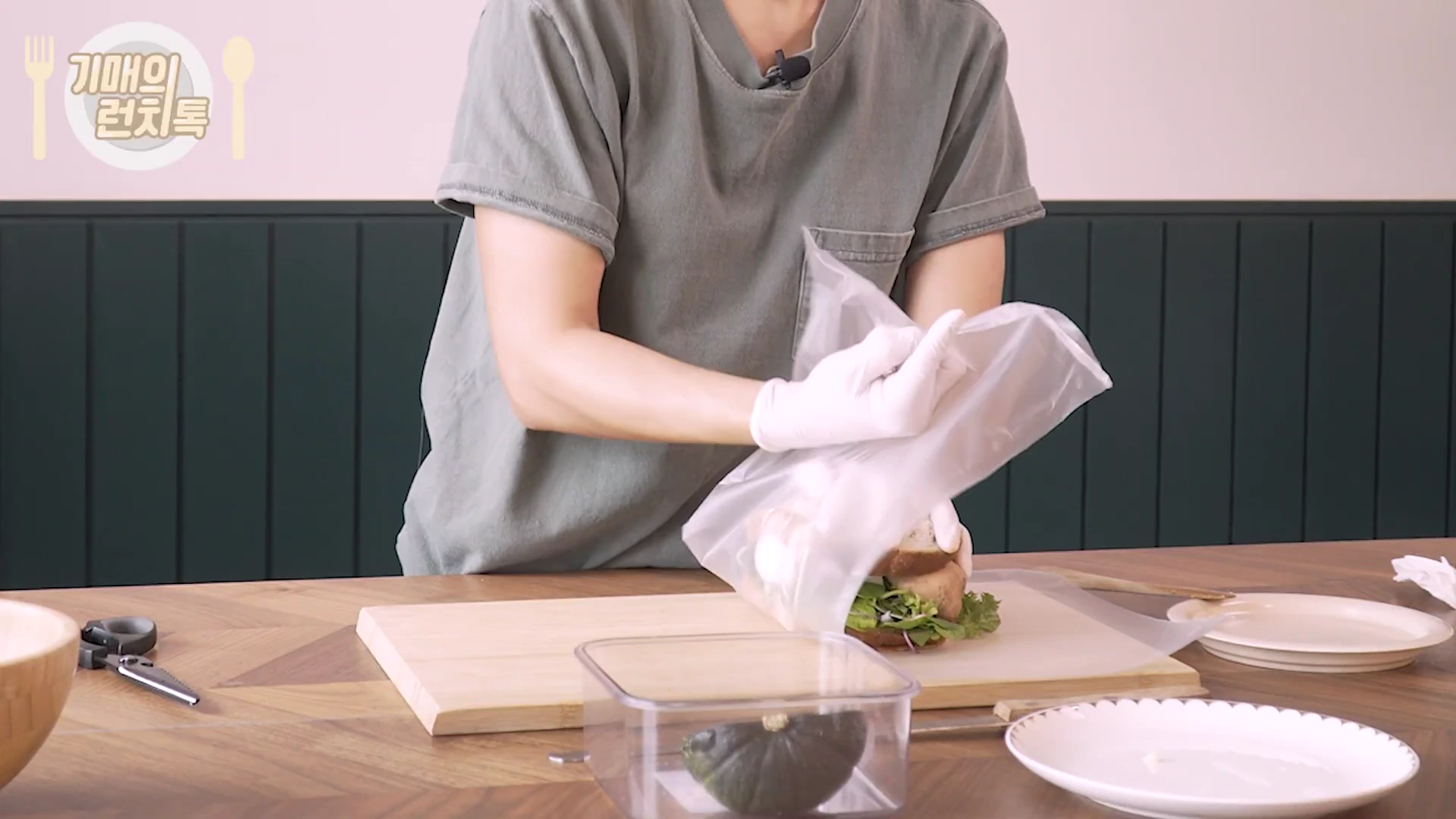흰색 위생 장갑을 낀 사람이 도마 위에서 샌드위치를 투명한 랩으로 꼼꼼하게 감싸고 있는 모습이다.