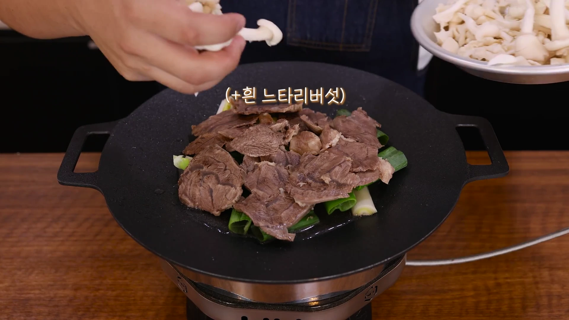 요리사의 손이 전골 냄비 위에 놓인 고기 위로 흰 느타리버섯을 올리고 있는 모습