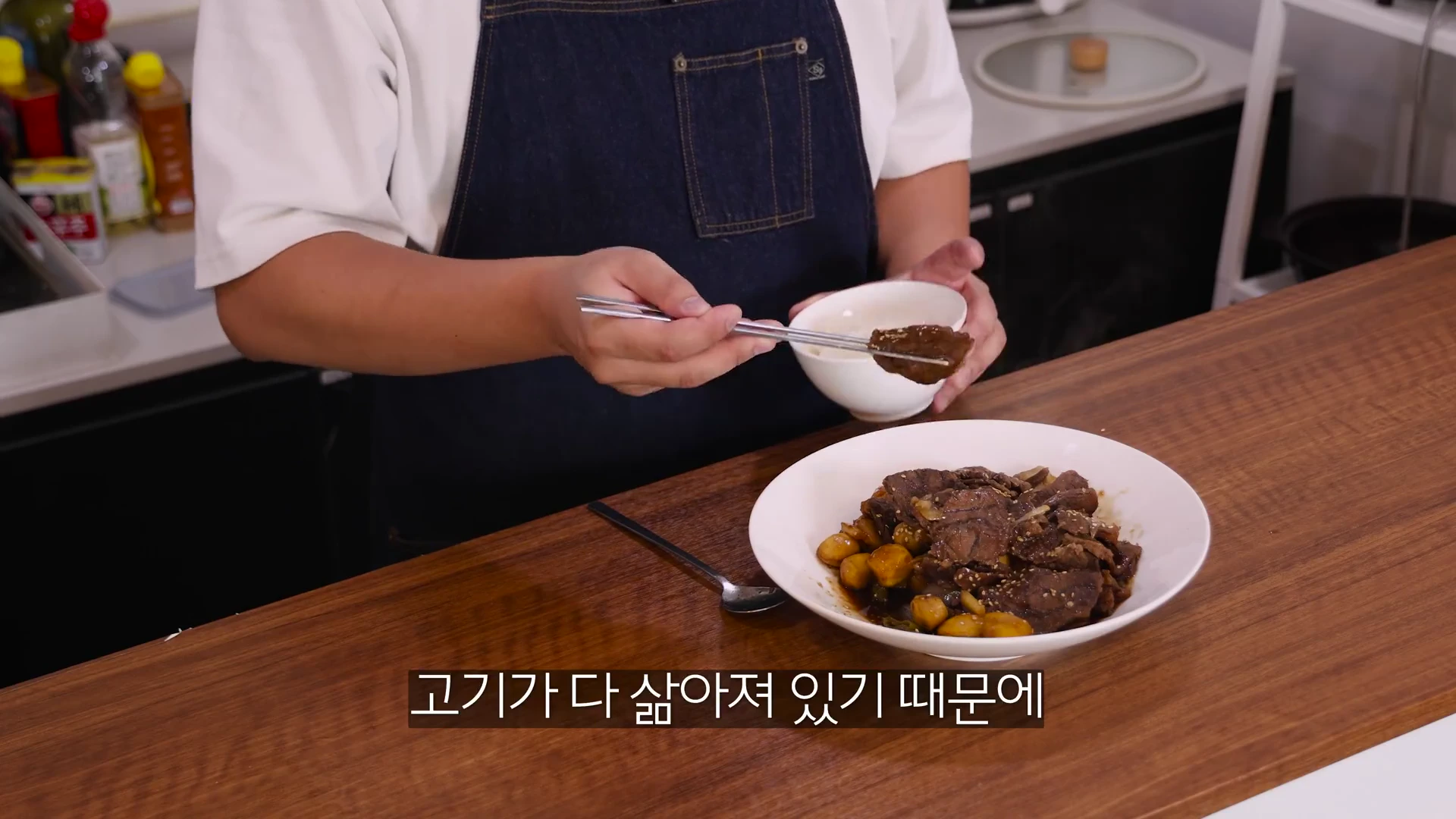 앞치마를 두른 요리사가 젓가락으로 갈비찜을 집어 들어 보여주고 있으며, 그 앞에는 밤과 고기가 담긴 그릇이 놓여 있습니다.