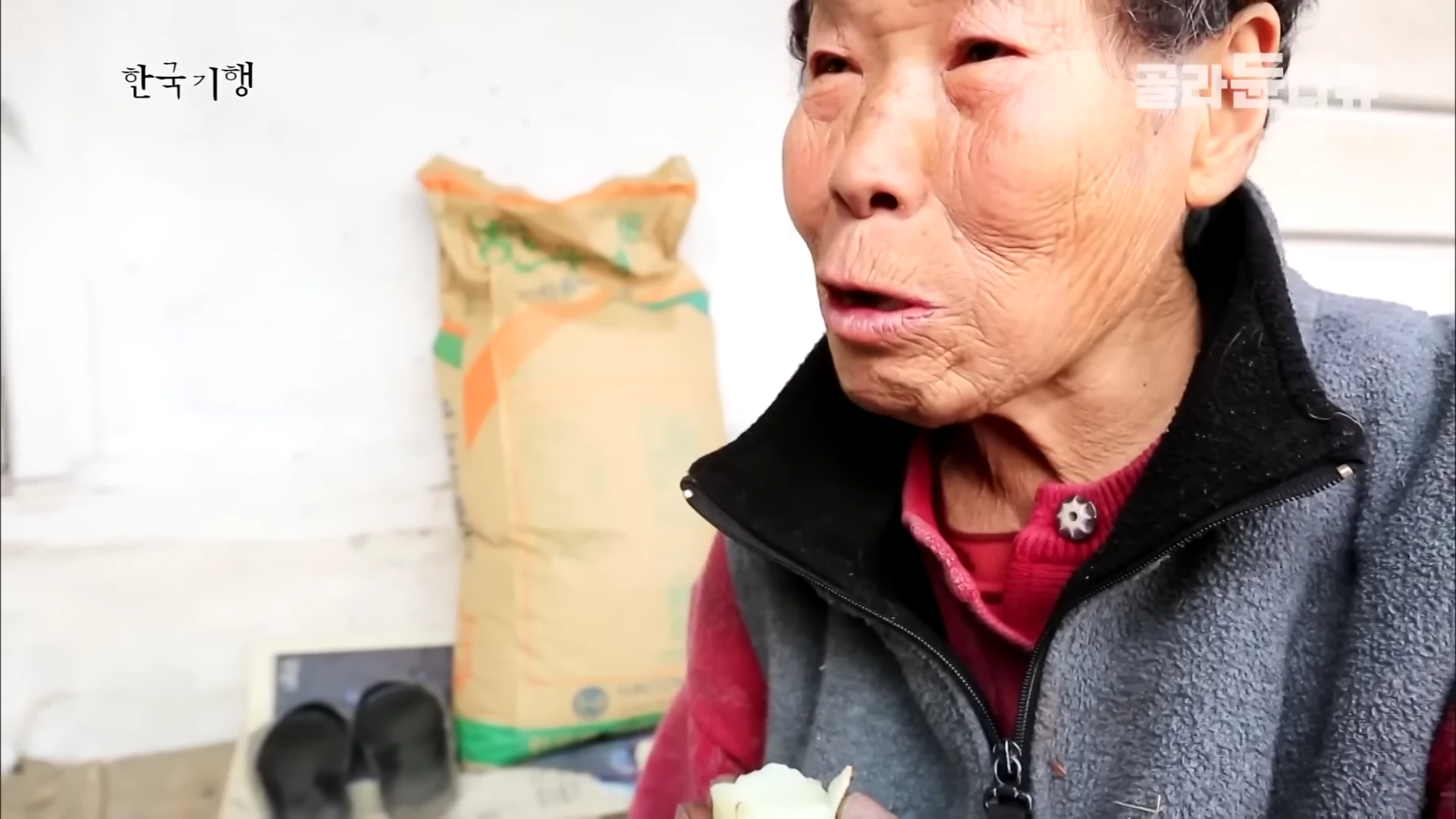 흰머리가 희끗한 노인 여성이 회색 조끼를 입고 숯불에 구운 감자를 먹으며 이야기하는 모습