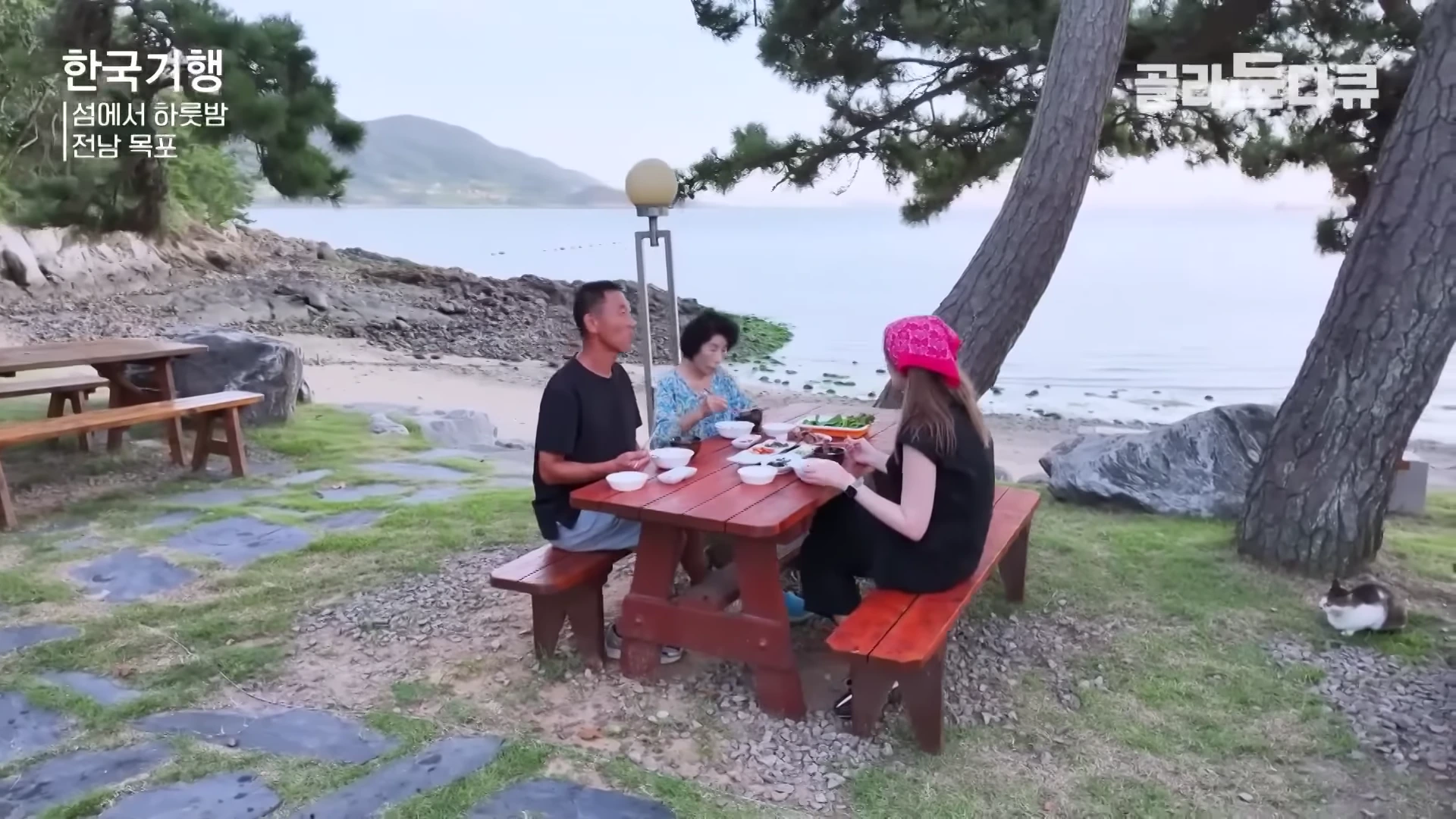바닷가에 놓인 나무 탁자에 세 사람이 둘러앉아 음식을 먹고 있고, 옆에는 고양이가 앉아 있는 모습