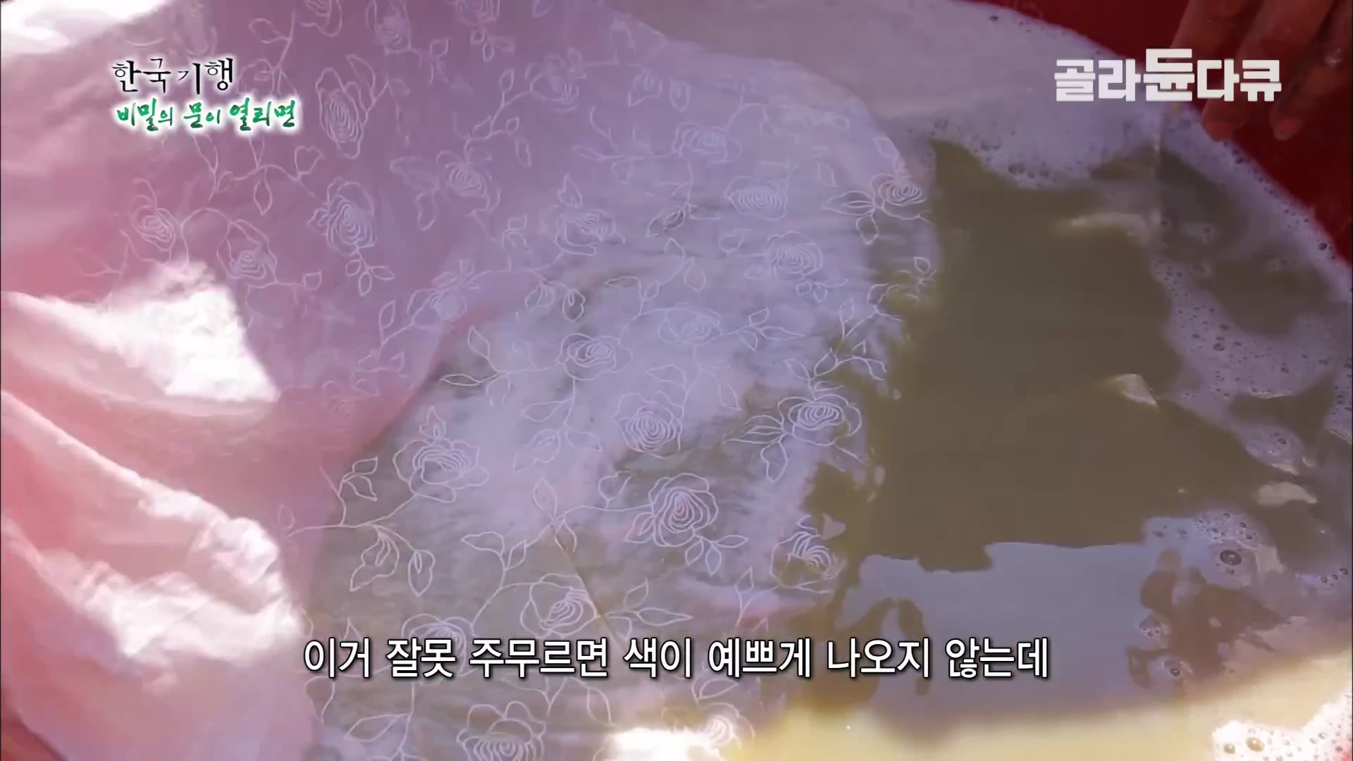 붉은색 대야 안에 든 갈색 염료 물에 장미 무늬가 있는 분홍색 천을 담가 정성스럽게 주무르고 있는 손의 모습.