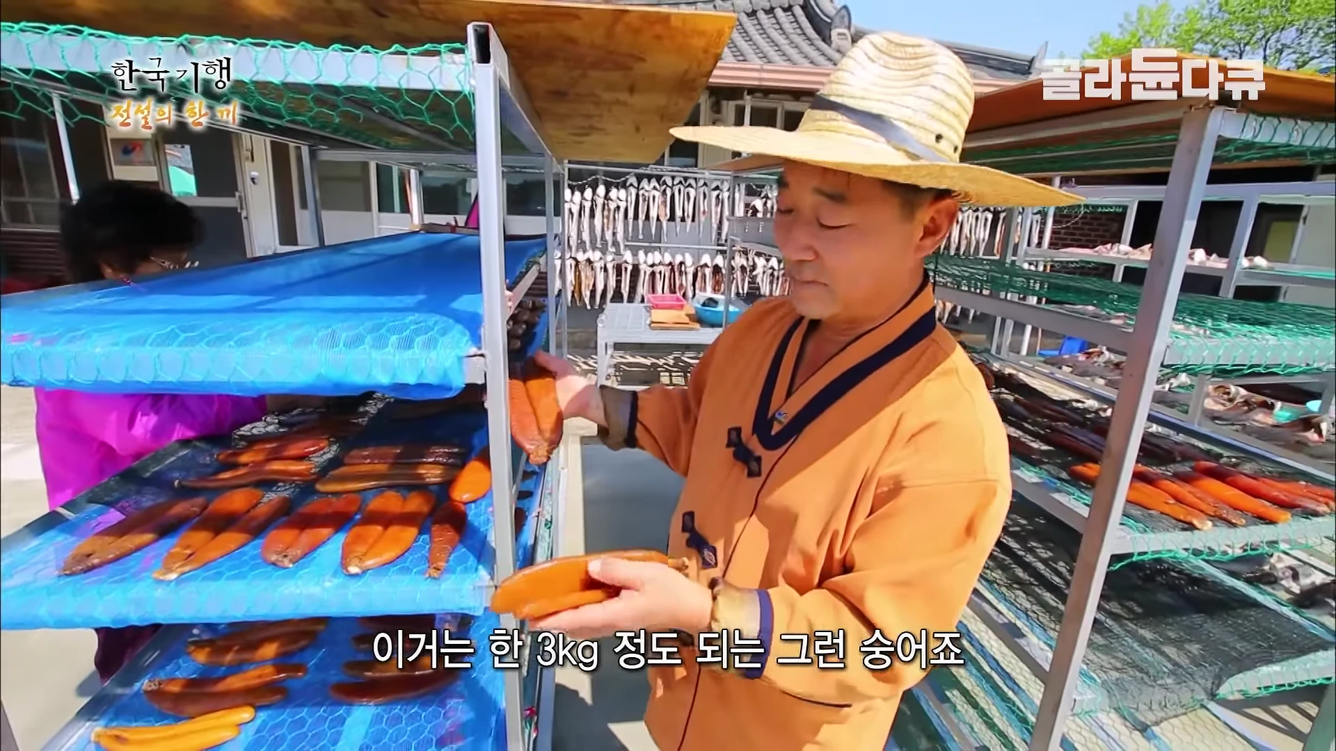 밀짚모자를 쓴 남성이 야외 건조대 앞에 서서 손에 든 어란을 살펴보고 있으며, 뒤쪽 건조대에는 줄지어 말리고 있는 생선들이 보인다.